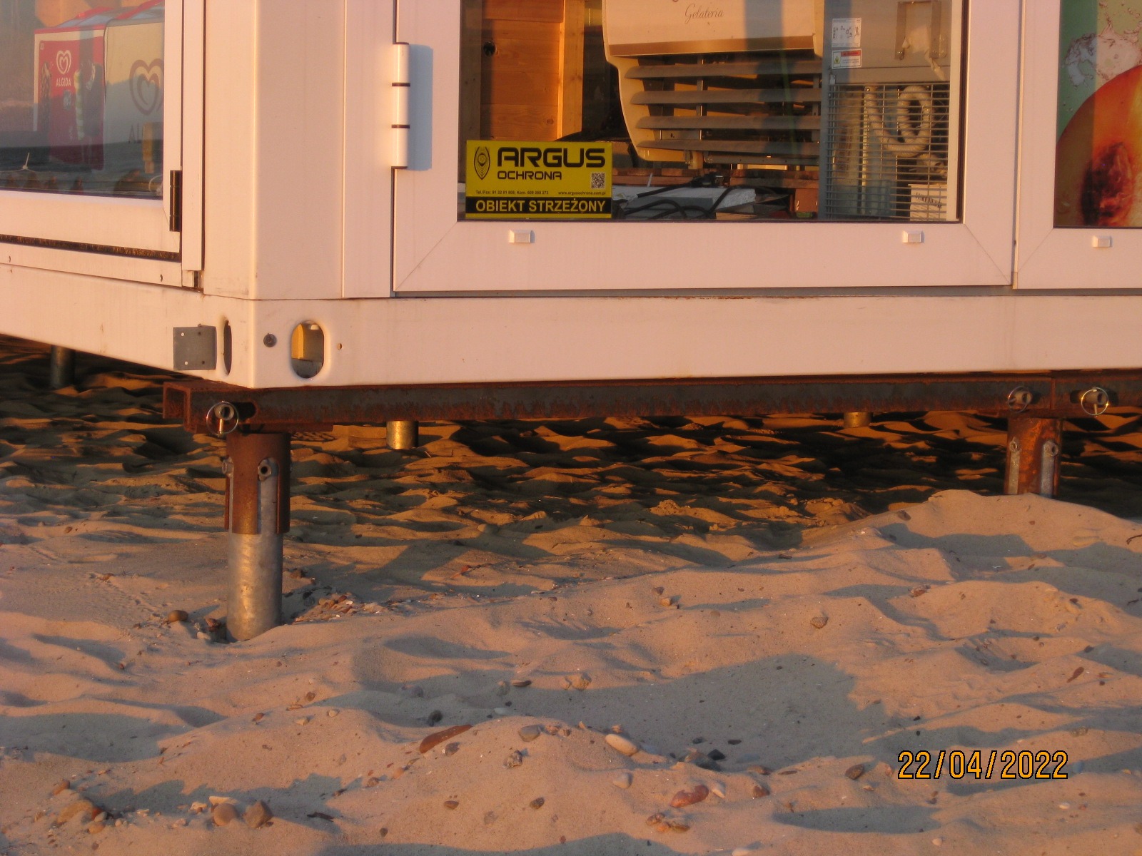 Biały, modułowy pawilon handlowy z metalową konstrukcją fundamentową, ustawiony na piaszczystej plaży w promieniach zachodzącego słońca. Widoczna tabliczka 'Obiekt Strzeżony'.