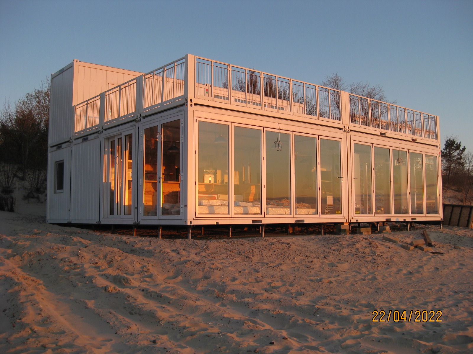 Nowoczesny, dwukondygnacyjny dom z kontenerów morskich, z balkonem na dachu, osadzony na piaszczystym podłożu. Duże przeszklenia odbijają zachodzące słońce.