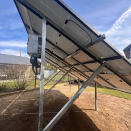 Widok od spodu na panele fotowoltaiczne zamontowane na metalowej konstrukcji, z widoczną skrzynką przyłączeniową i przewodami, w tle ogród z trawnikiem i budynkiem.