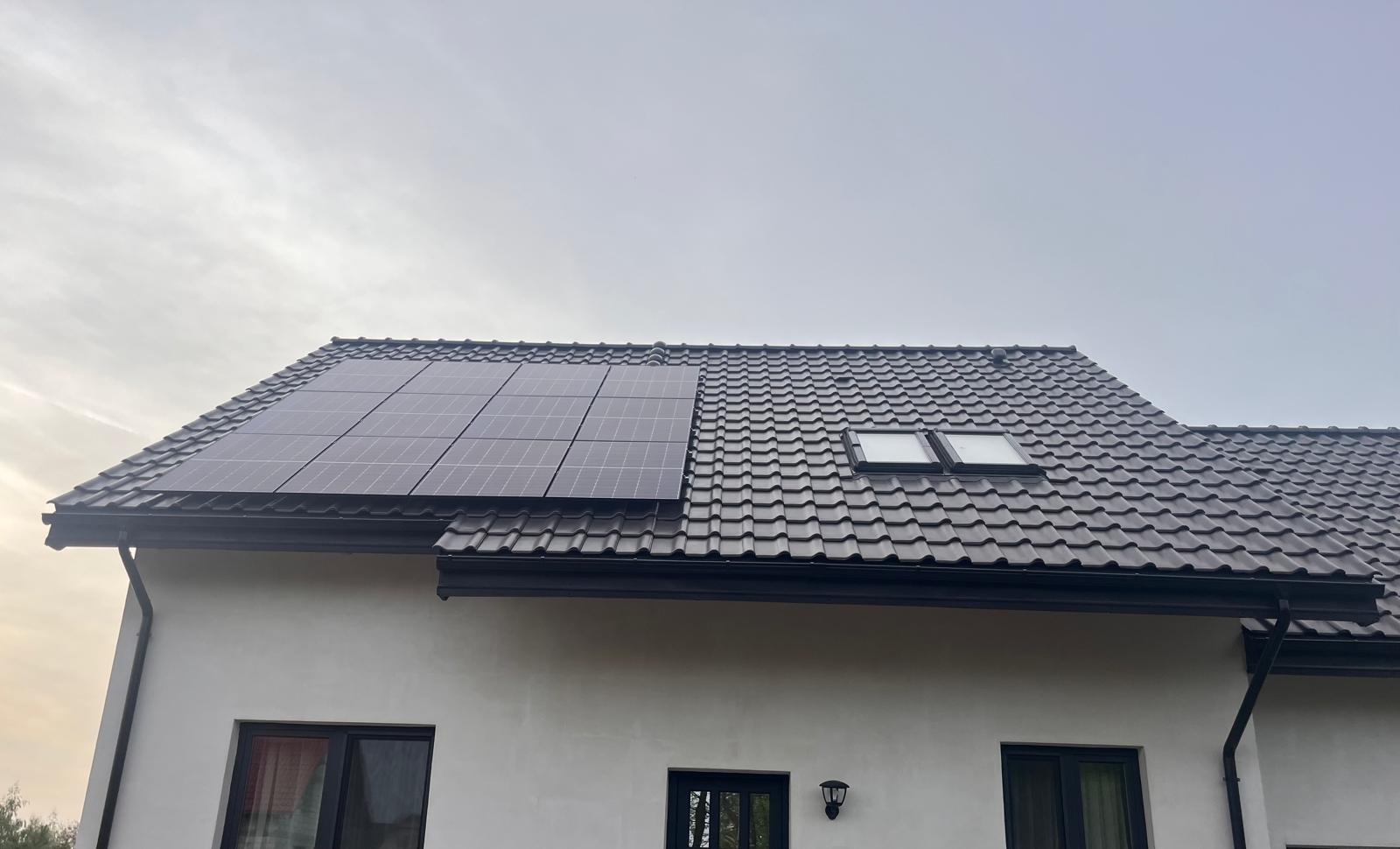 Panele słoneczne na dachu domu z ciemną dachówką i dwoma oknami dachowymi, widoczne fragmenty elewacji i okien.