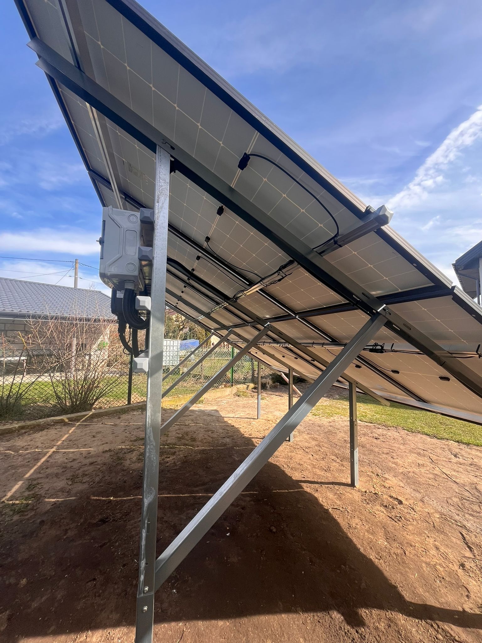 Widok od spodu na panele fotowoltaiczne zamontowane na metalowej konstrukcji, z widoczną skrzynką przyłączeniową i przewodami, w tle ogród z trawnikiem i budynkiem.