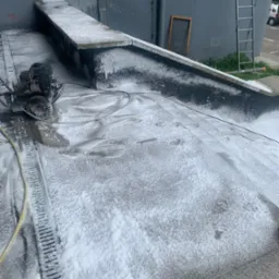 Dach pokryty pianą czyszczącą, widoczna maszyna czyszcząca z silnikiem spalinowym i żółtym wężem, drabina oparta o szarą ścianę budynku w tle.