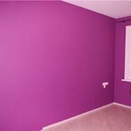 MatBut - Pusty pokój z fioletowymi ścianami i beżową podłogą. Białe okno z żaluzjami i grzejnik pod nim. Widoczny kontakt elektryczny na ścianie.