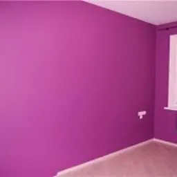 Pusty pokój z fioletowymi ścianami i beżową podłogą. Białe okno z żaluzjami i grzejnik pod nim. Widoczny kontakt elektryczny na ścianie.