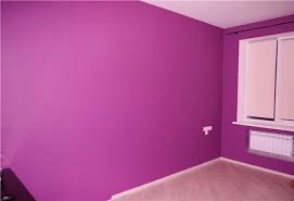 Pusty pokój z fioletowymi ścianami i beżową podłogą. Białe okno z żaluzjami i grzejnik pod nim. Widoczny kontakt elektryczny na ścianie.