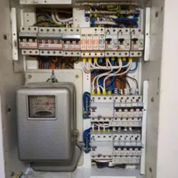 Otwarta skrzynka elektryczna z licznikiem i zabezpieczeniami, widoczne przewody i elementy instalacji. Kable uporządkowane, ale widać ślady użytkowania.