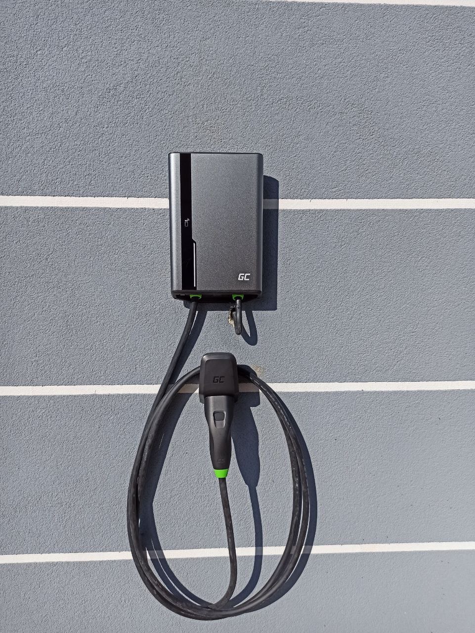 Szara stacja ładowania pojazdów elektrycznych GC zamontowana na szarej ścianie z poziomymi pasami, kabel z wtyczką zwisa w pętli.