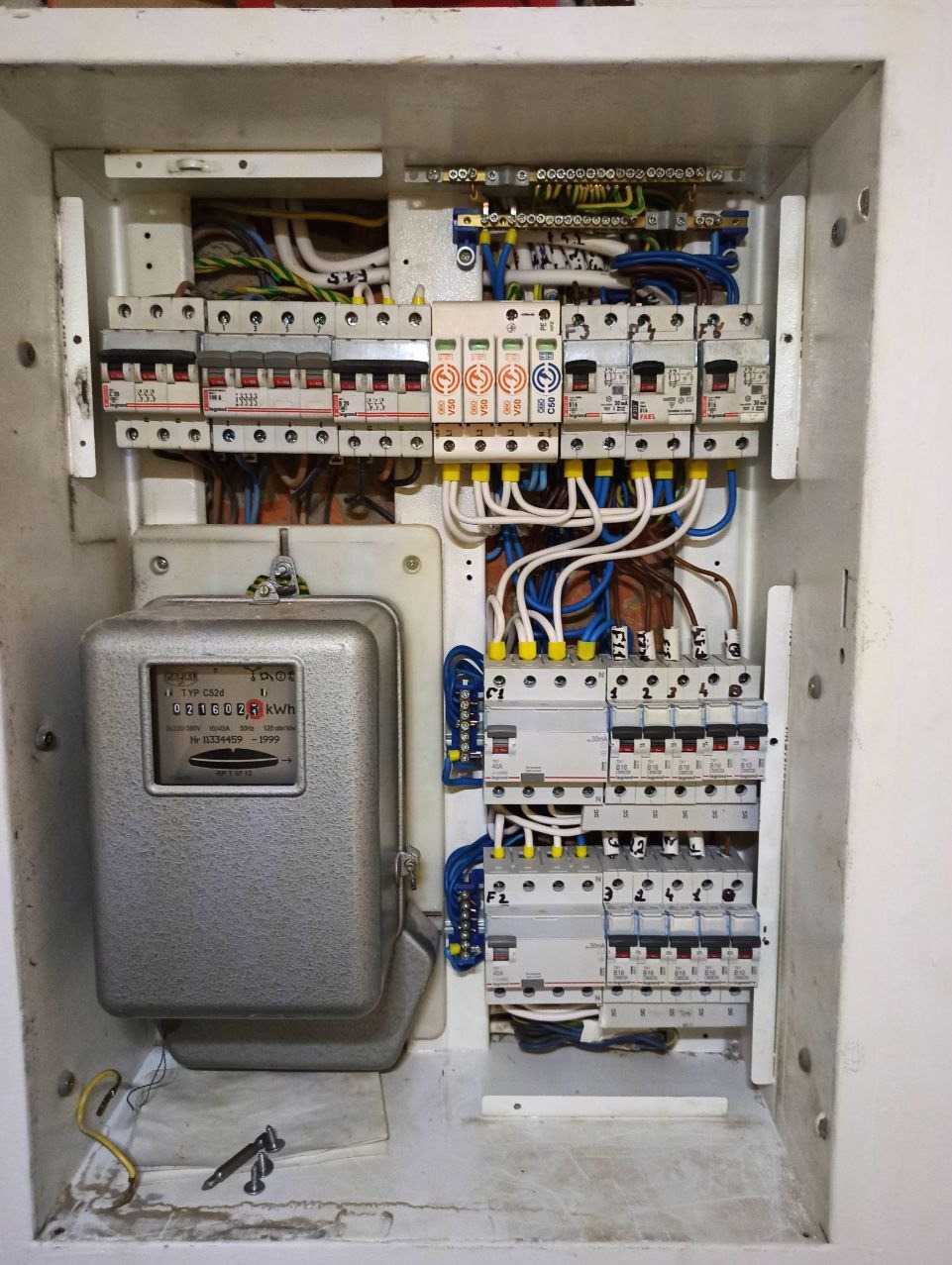 Otwarta skrzynka elektryczna z licznikiem i zabezpieczeniami, widoczne przewody i elementy instalacji. Kable uporządkowane, ale widać ślady użytkowania.