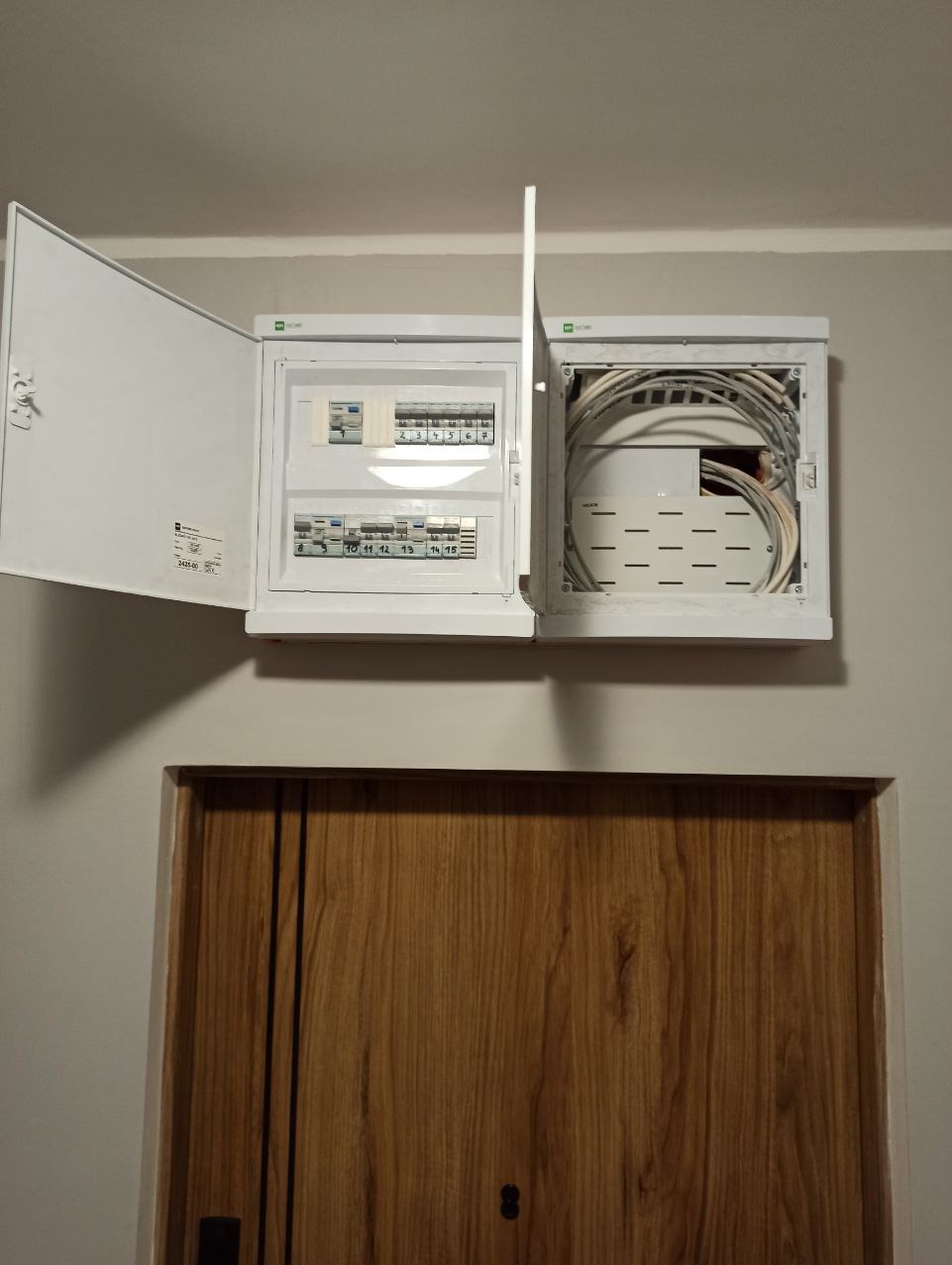 Otwarta, biała skrzynka rozdzielcza z bezpiecznikami i kablami, zamontowana nad drewnianymi drzwiami w jasnym pomieszczeniu. Widoczne numerowane bezpieczniki i kable wewnątrz.