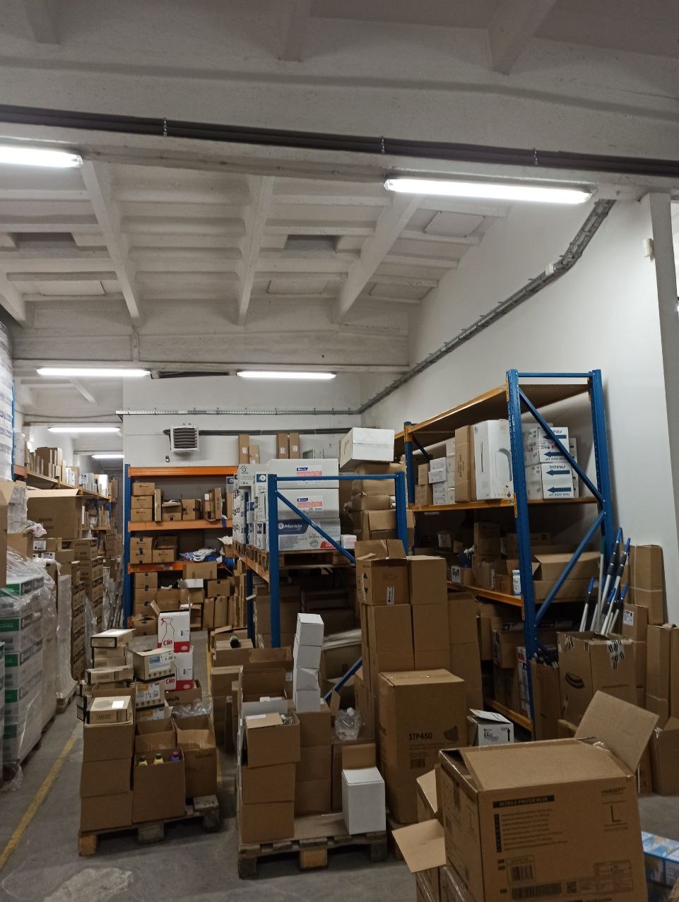 Wnętrze magazynu z regałami i kartonami, oświetlone lampami sufitowymi. Widoczne instalacje pod sufitem. Perspektywa z dołu, ukazująca dużą przestrzeń składowania.