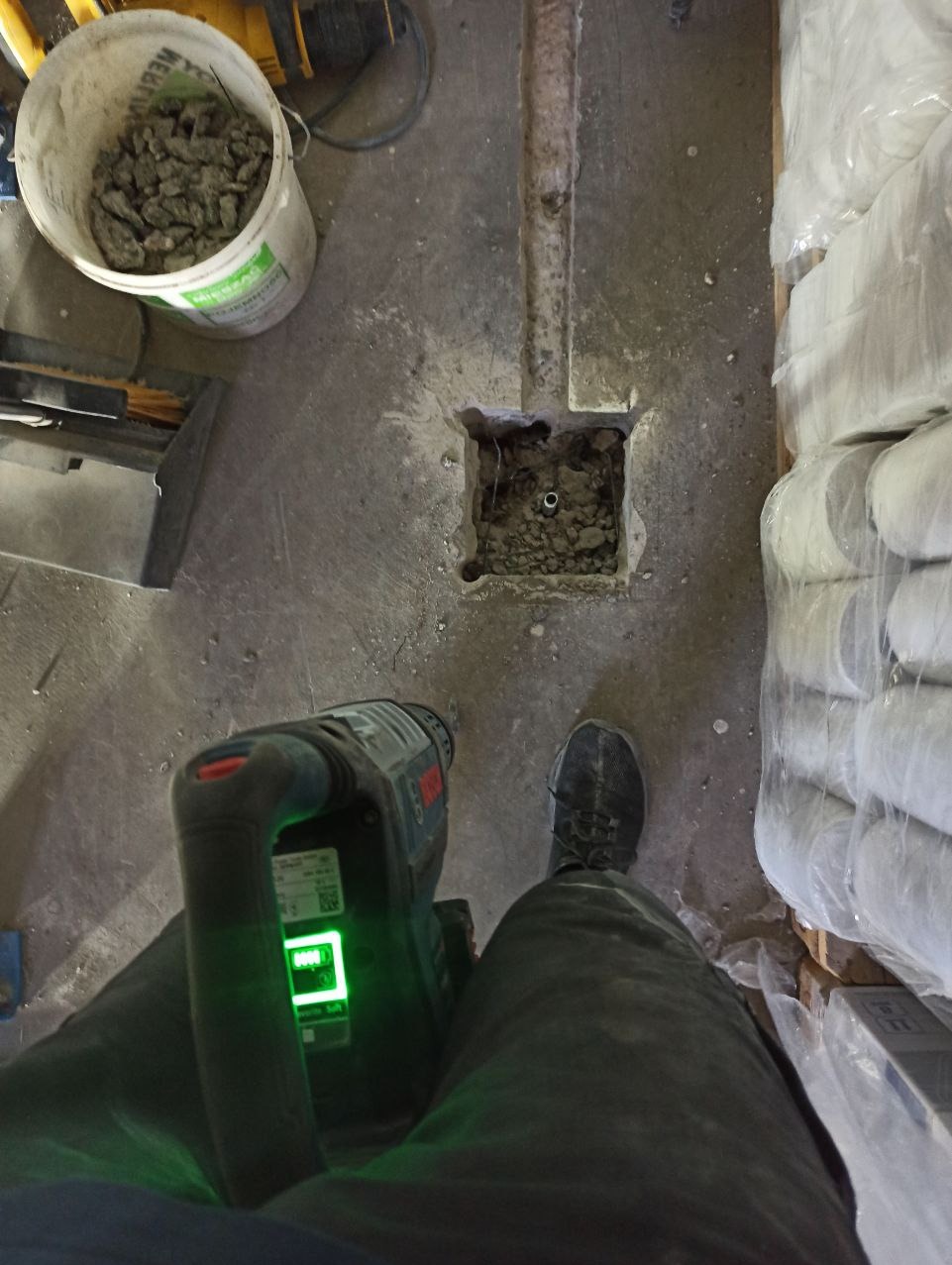 Wykuty otwór w podłodze z widoczną rurką, obok leży wiertarka z włączonym laserem, wiadro z gruzem i worki z zaprawą. Przygotowanie do prac instalacyjnych.