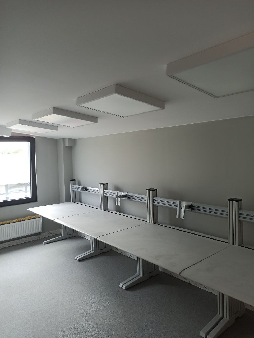 Nowoczesne biuro z podwieszanymi, kwadratowymi lampami LED, szarymi ścianami i długim stołem konferencyjnym z metalową konstrukcją wsporczą.