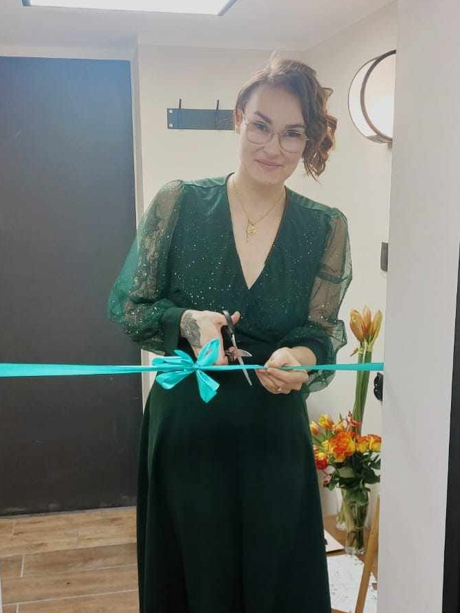 Uśmiechnięta kobieta w zielonej sukience przecina turkusową wstęgę nożyczkami podczas otwarcia gabinetu