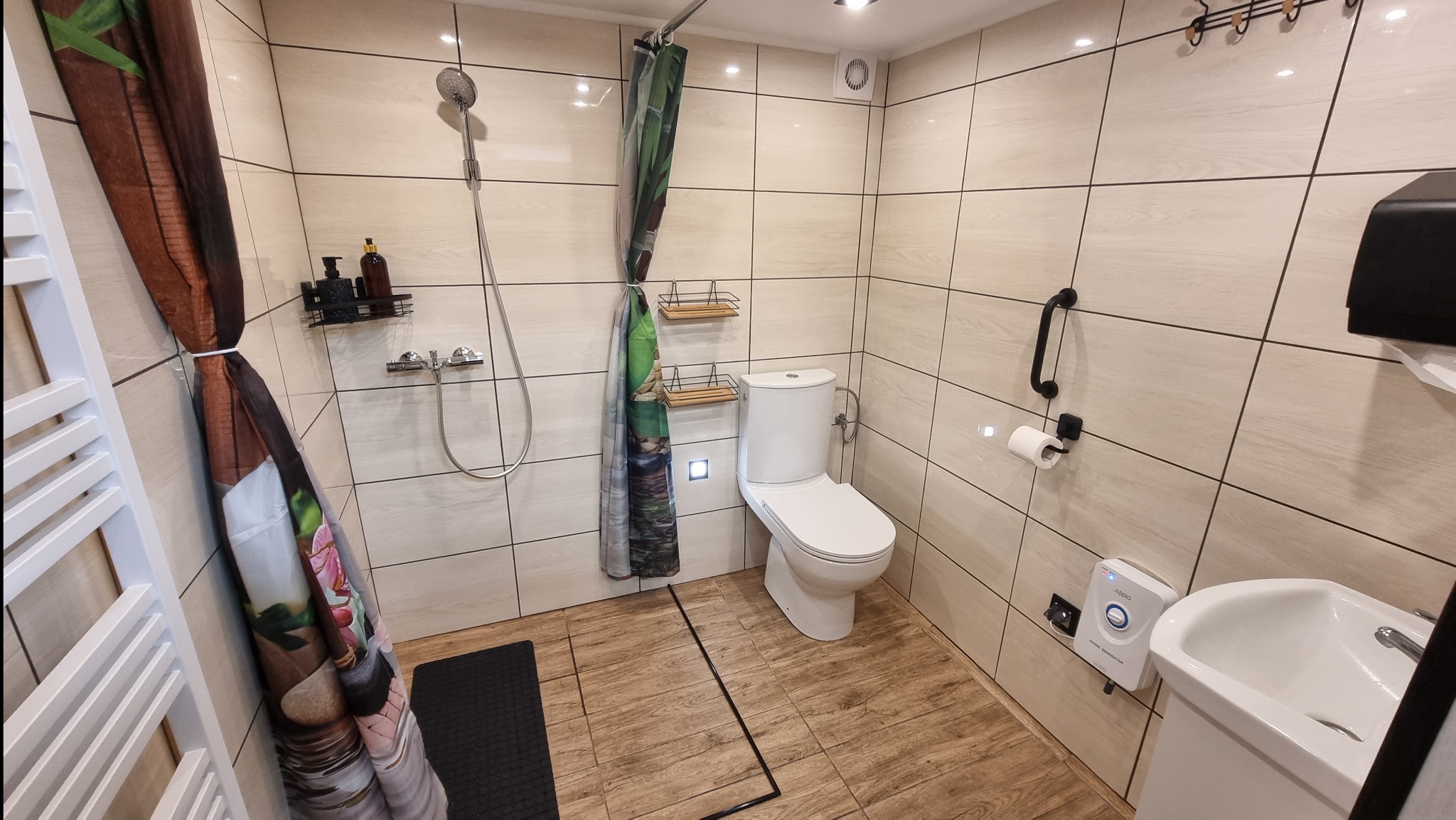 Łazienka z prysznicem i toaletą w Pieńsku, wykończona w jasnych płytkach imitujących drewno, z kolorową zasłoną prysznicową i dodatkowymi półkami.