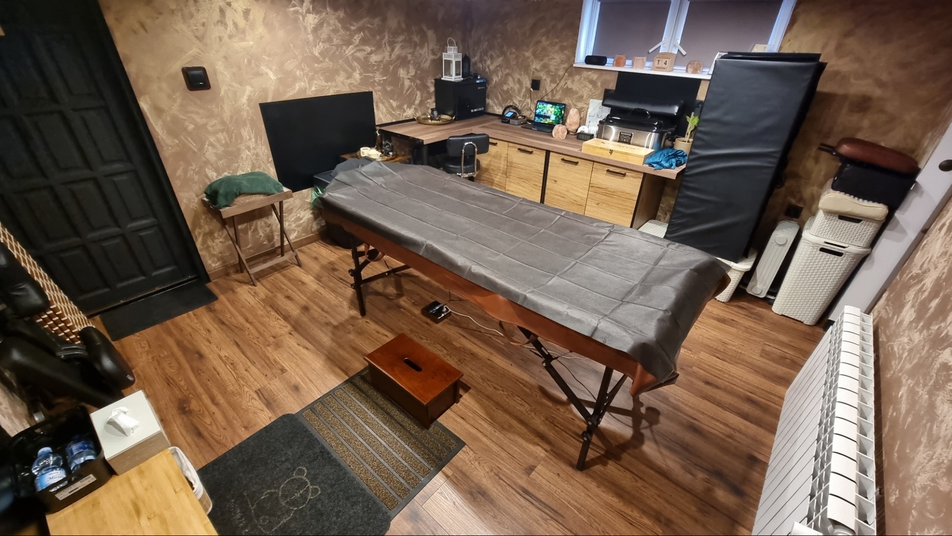 Przygotowane stanowisko do zabiegów z rozłożonym stołem pokrytym szarą narzutą, drewnianą podestem i dywanikiem z motywem misia, w tle biurko z laptopem i akcesoriami.