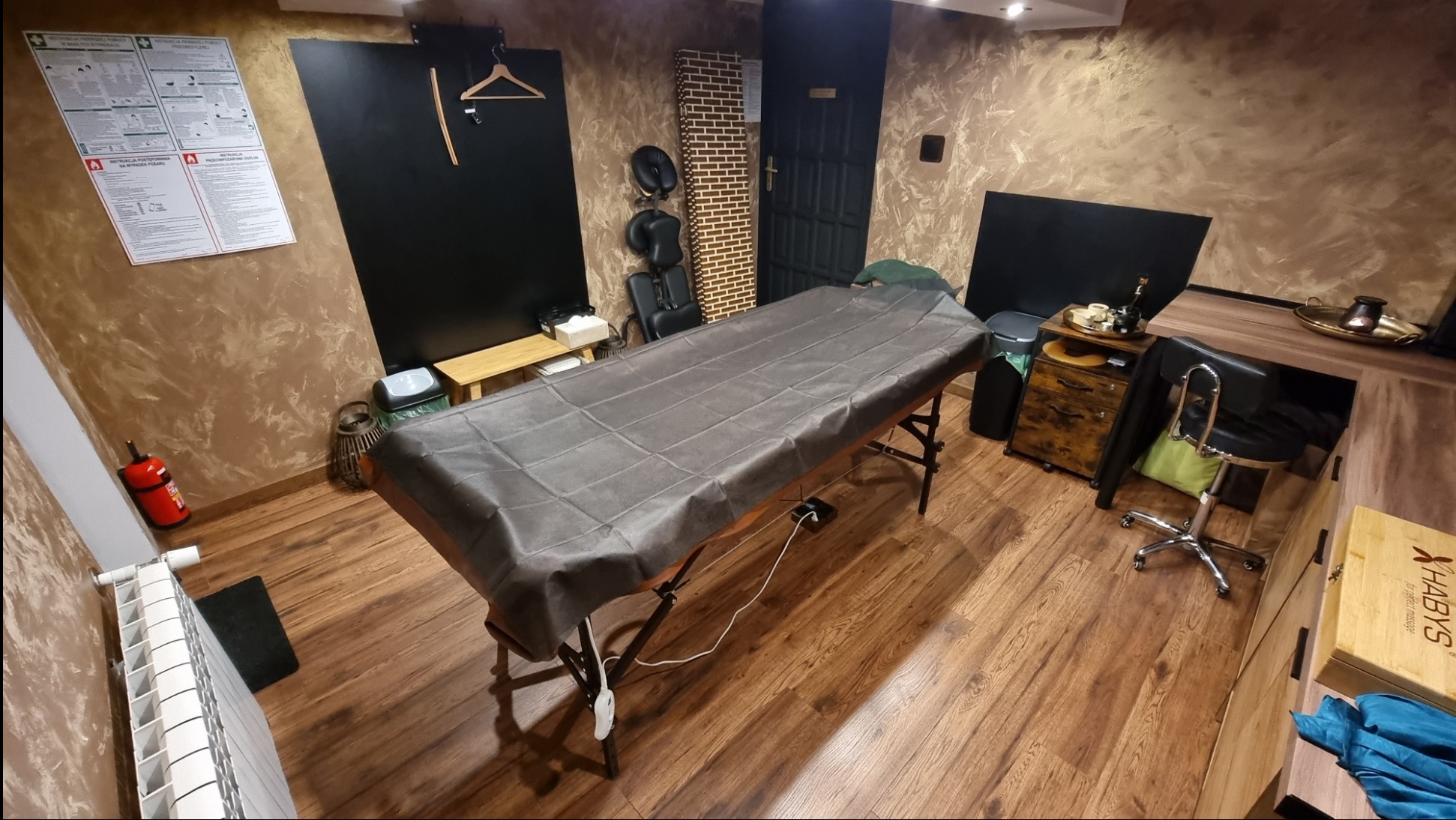 Przygotowane stanowisko do masażu: stół przykryty szarym pokrowcem, w tle krzesło do masażu, instrukcja pierwszej pomocy i gaśnica w rogu. Widoczne elementy wystroju wnętrza.