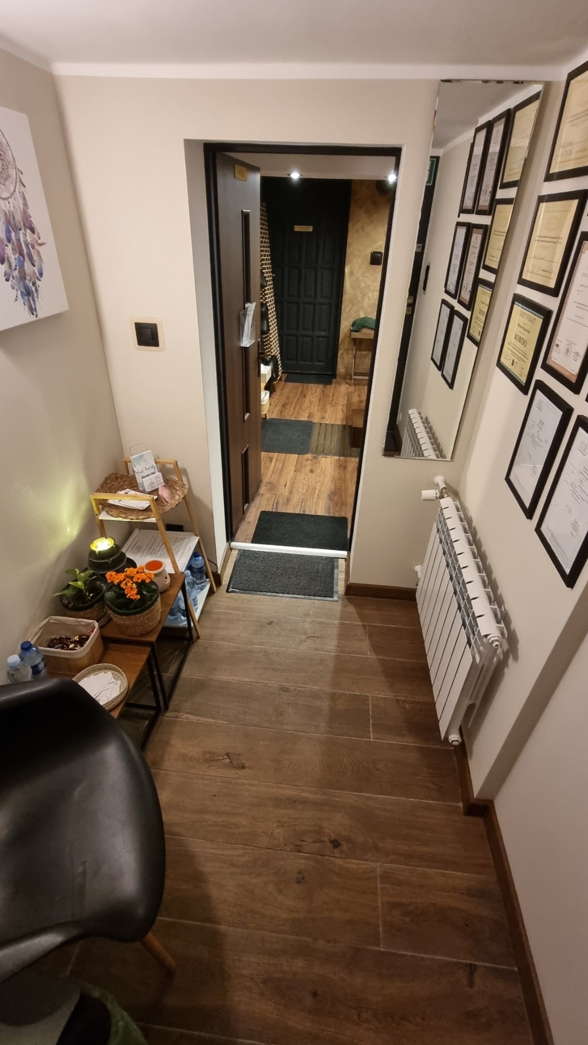 Wnętrze salonu z drewnianą podłogą, widok na otwarte drzwi do gabinetu z czarnymi elementami, dekoracje na ścianie, w tym lustro z certyfikatami i obraz z łapaczem snów.