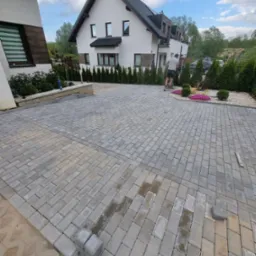 Ułożona kostka brukowa na podjeździe przed nowoczesnym domem jednorodzinnym; widoczne mokre plamy na powierzchni, sugerujące świeże prace, oraz pracownik w oddali.