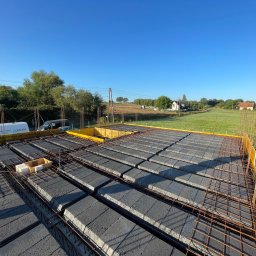 BALEN Inżynieria Budowlana Dawid Balicki - Zbrojenie stropu Teriva z szalunkiem w żółtej formie, widok z góry na tle krajobrazu z domami i błękitnym niebem. Na pierwszym planie skrzynka z elementami.