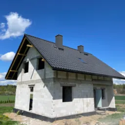 Niedokończony dom z białego betonu komórkowego z czarnym dachem, kominami i oknem dachowym. Widoczne elementy drewniane więźby dachowej. W tle błękitne niebo i zieleń.