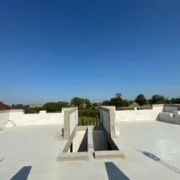 Niedokończony strop budynku z białych bloczków, z widocznymi otworami i betonowymi elementami, w tle zielony krajobraz i błękitne niebo. Budowa w toku.