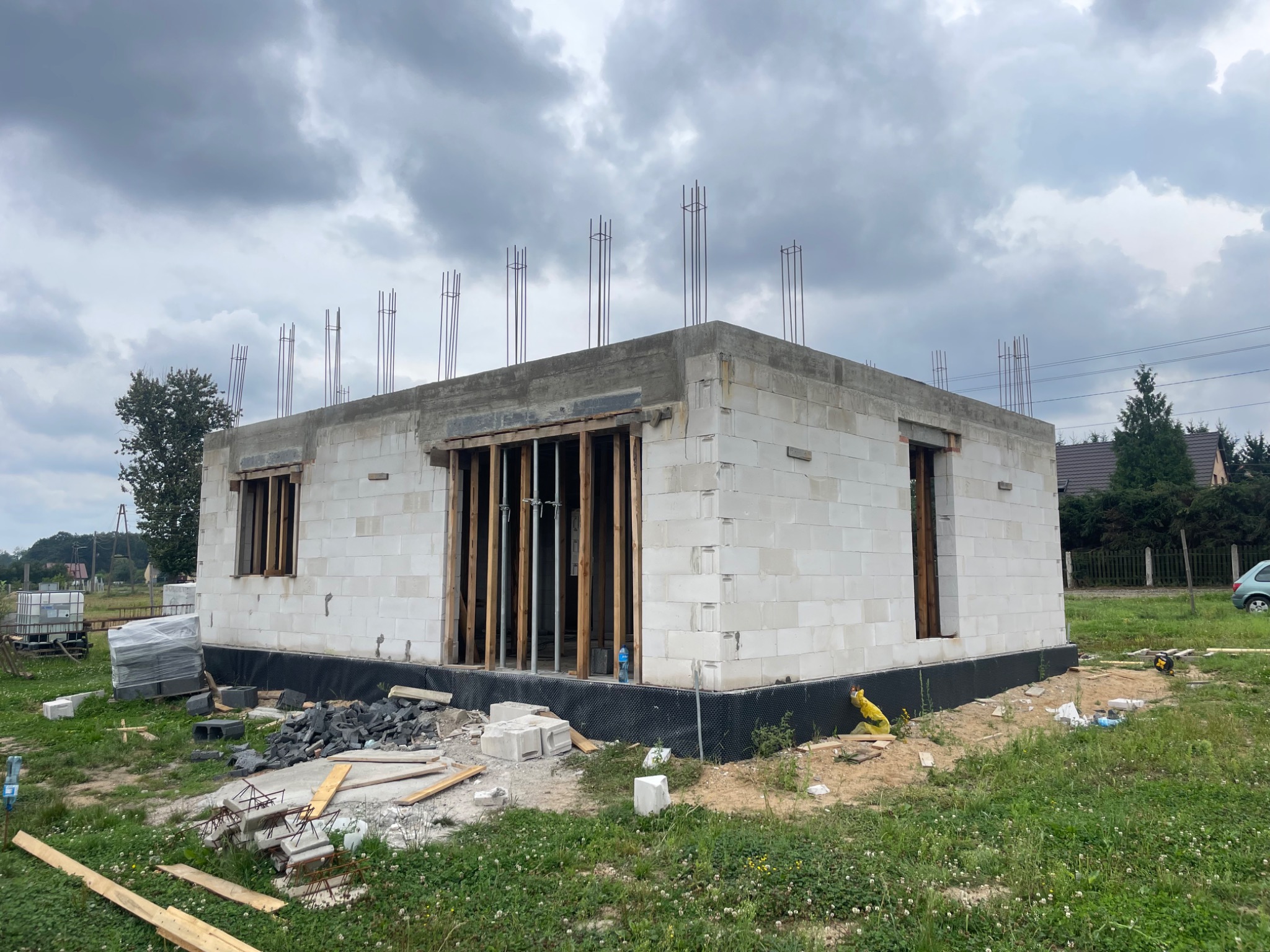 Niedokończony budynek z białych bloczków, z widoczną izolacją fundamentów i wystającymi prętami zbrojeniowymi, otoczony zielenią i materiałami budowlanymi.