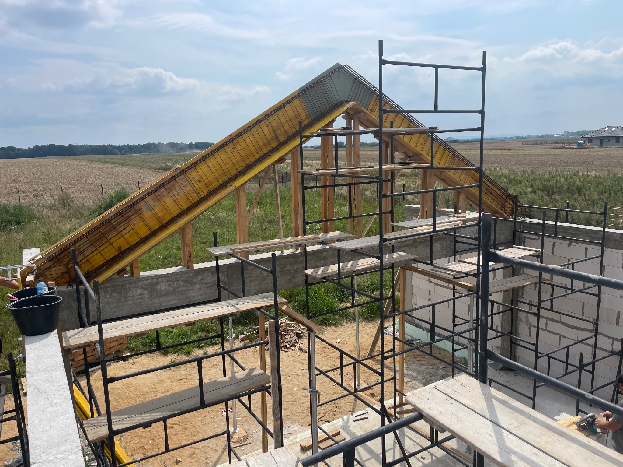 Wznoszenie konstrukcji dachu z szalunkiem i zbrojeniem na nowo budowanym domu z bloczków, widok z rusztowania na otwarty krajobraz.