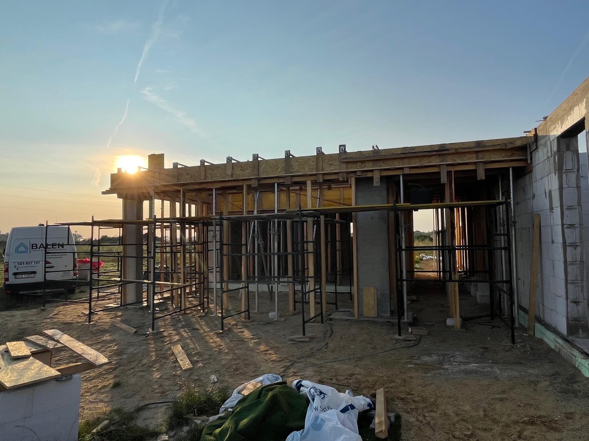 Wznoszenie ścian domu z bloczków betonowych, widoczne szalunki i rusztowania, w tle zachodzące słońce i firmowy van.