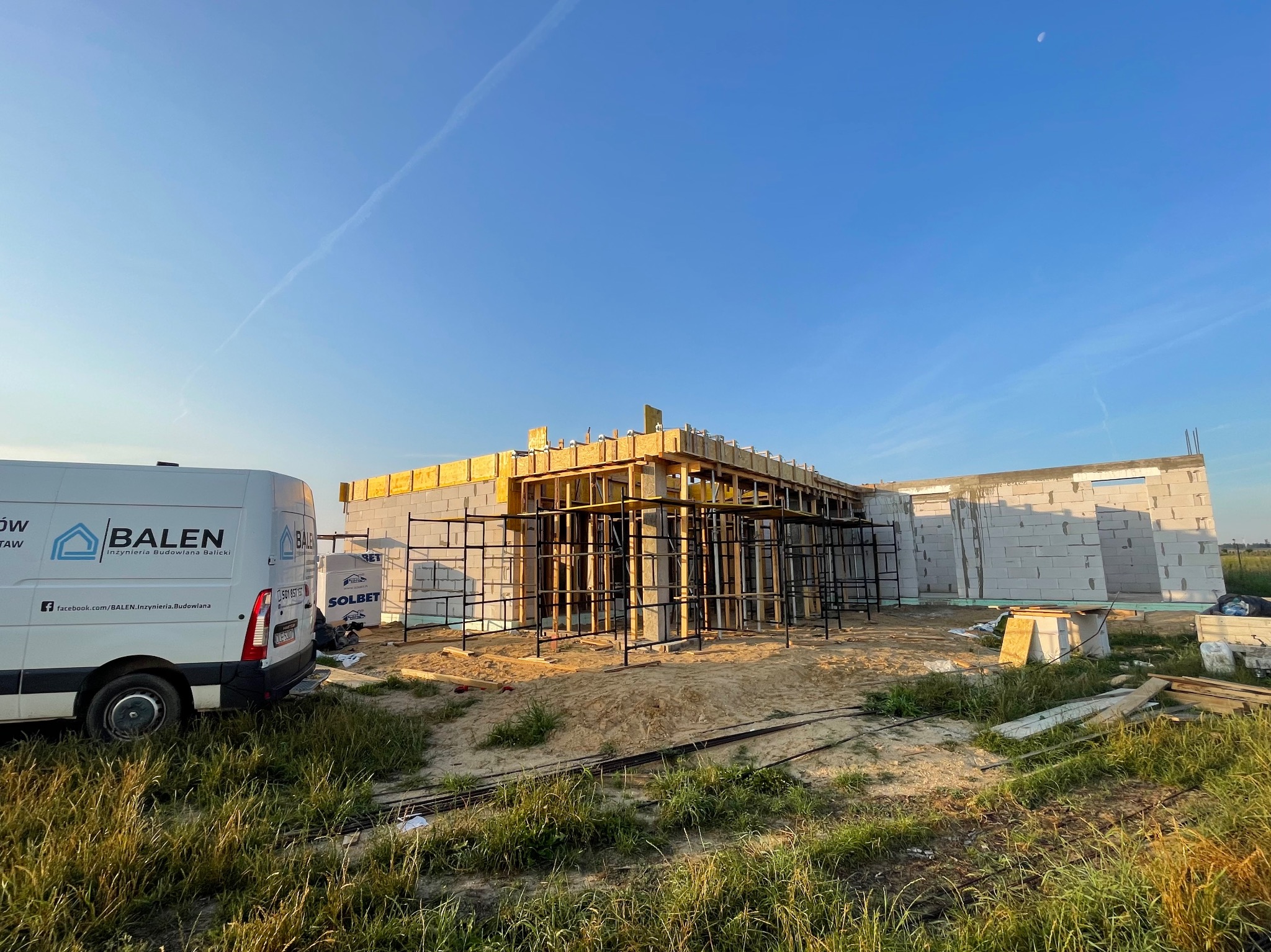 Budowa domu murowanego w trakcie wznoszenia ścian z betonu komórkowego Solbet; widoczne rusztowania, drewniane szalunki i firmowy bus BALEN w tle pod błękitnym niebem.