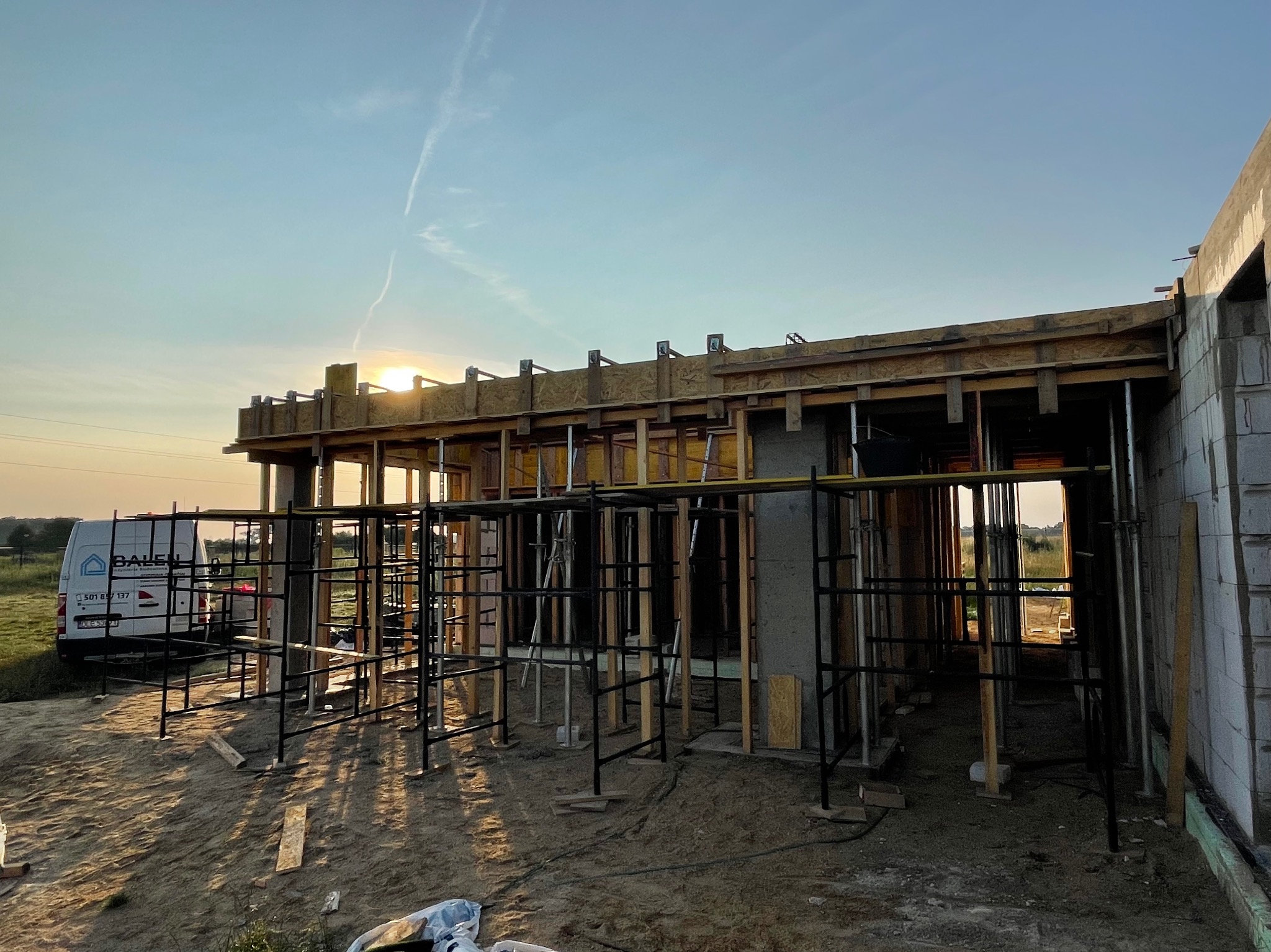 Wczesna faza budowy domu: widoczne szalunki, rusztowania i częściowo wymurowane ściany z betonu komórkowego w promieniach zachodzącego słońca. Na drugim planie zaparkowany biały bus firmy budowlanej.