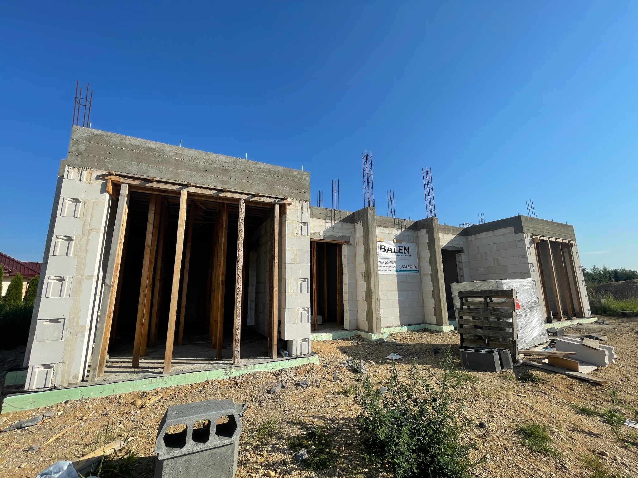 Wznoszenie ścian parterowego domu z betonu komórkowego, widoczne zbrojenie, szalunki okienne i paleta z materiałami budowlanymi w słoneczny dzień.