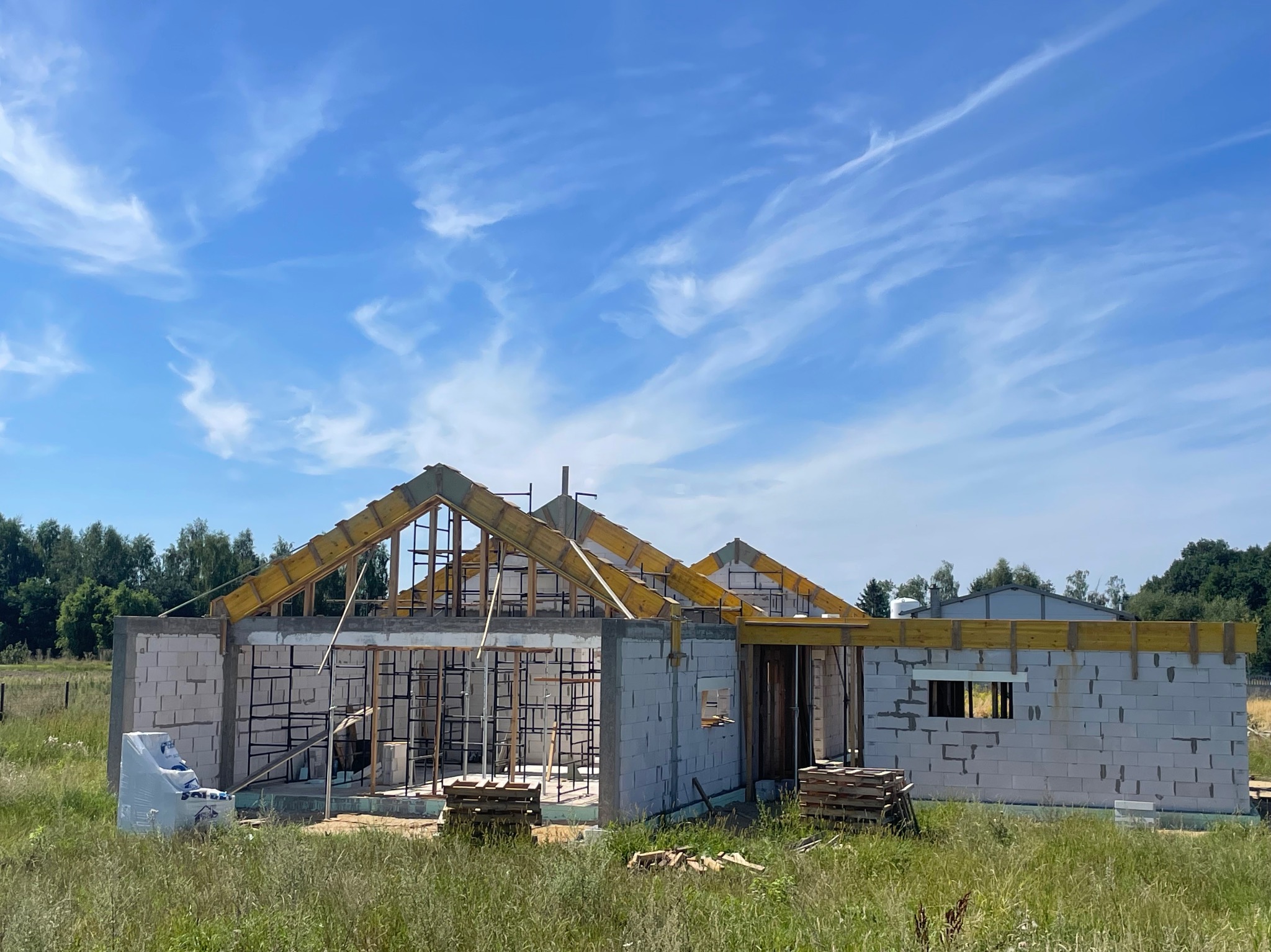Niedokończony budynek z białych bloczków, widoczne drewniane szalunki dachu, na pierwszym planie trawa i materiały budowlane w folii.