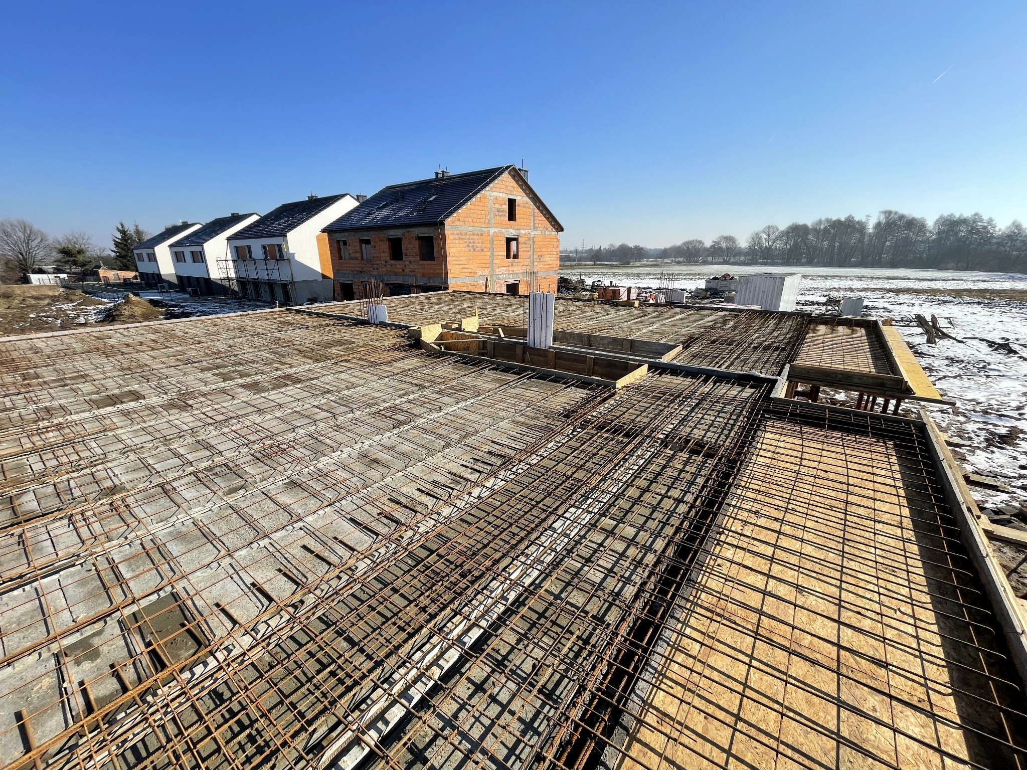 Zbrojenie stropu domu jednorodzinnego na tle budynków w zabudowie szeregowej i zaśnieżonego pola. Widok z góry na siatkę z prętów i szalunki.