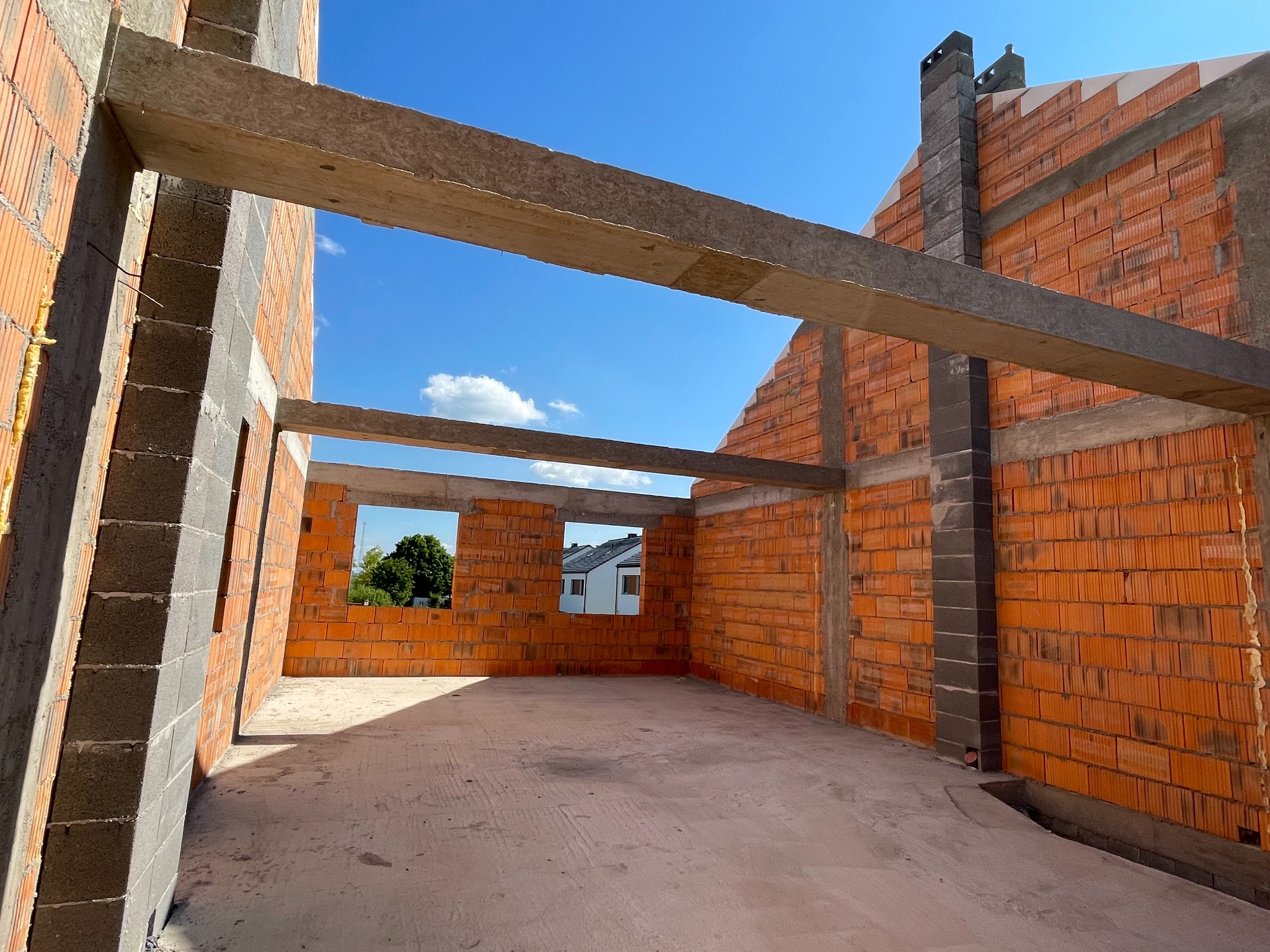 Wnętrze domu w stanie surowym otwartym: pomarańczowe ściany z pustaków, betonowe nadproża, widok na błękitne niebo przez okna. Kominek z ciemnej cegły.