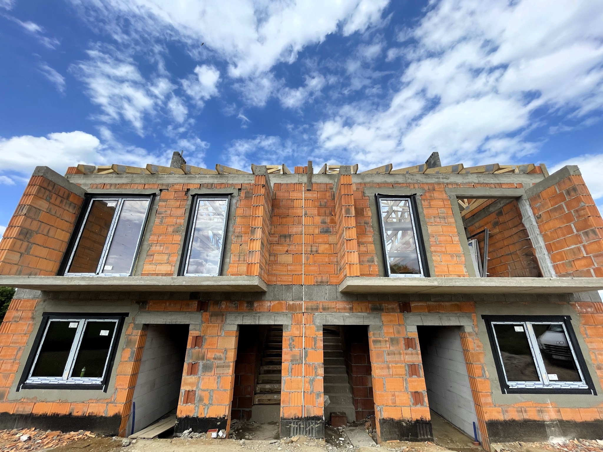 Wznoszenie bliźniaka z czerwonej cegły dziurawki, widok z dołu na niezadaszony budynek z zamontowanymi oknami, na tle błękitnego nieba z chmurami.