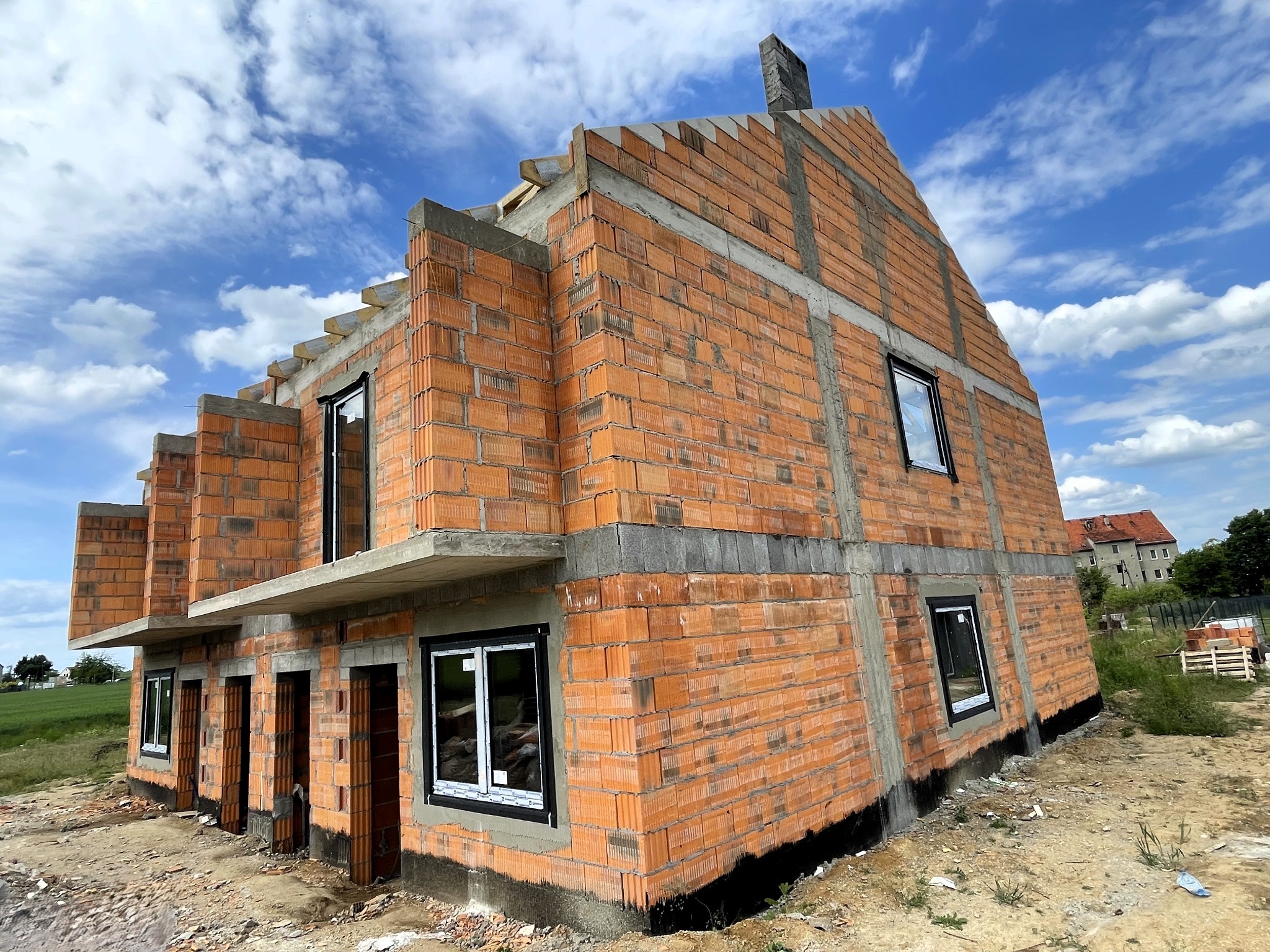 Niedokończony dom z czerwonej cegły w słoneczny dzień. Widoczne okna z czarnymi ramami i szare betonowe nadproża. W tle błękitne niebo i chmury.