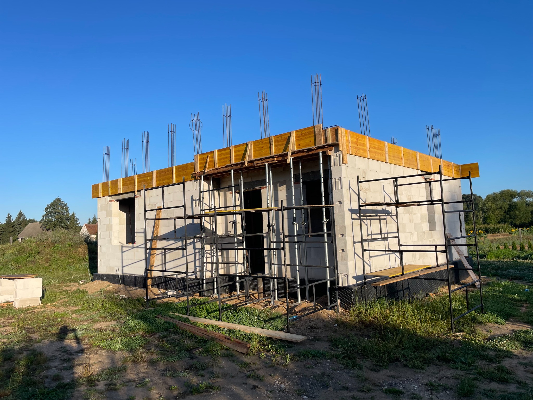 Surowy stan domu z betonu komórkowego, z widocznymi rusztowaniami i zbrojeniami, w otoczeniu zieleni i błękitnego nieba, w trakcie budowy.