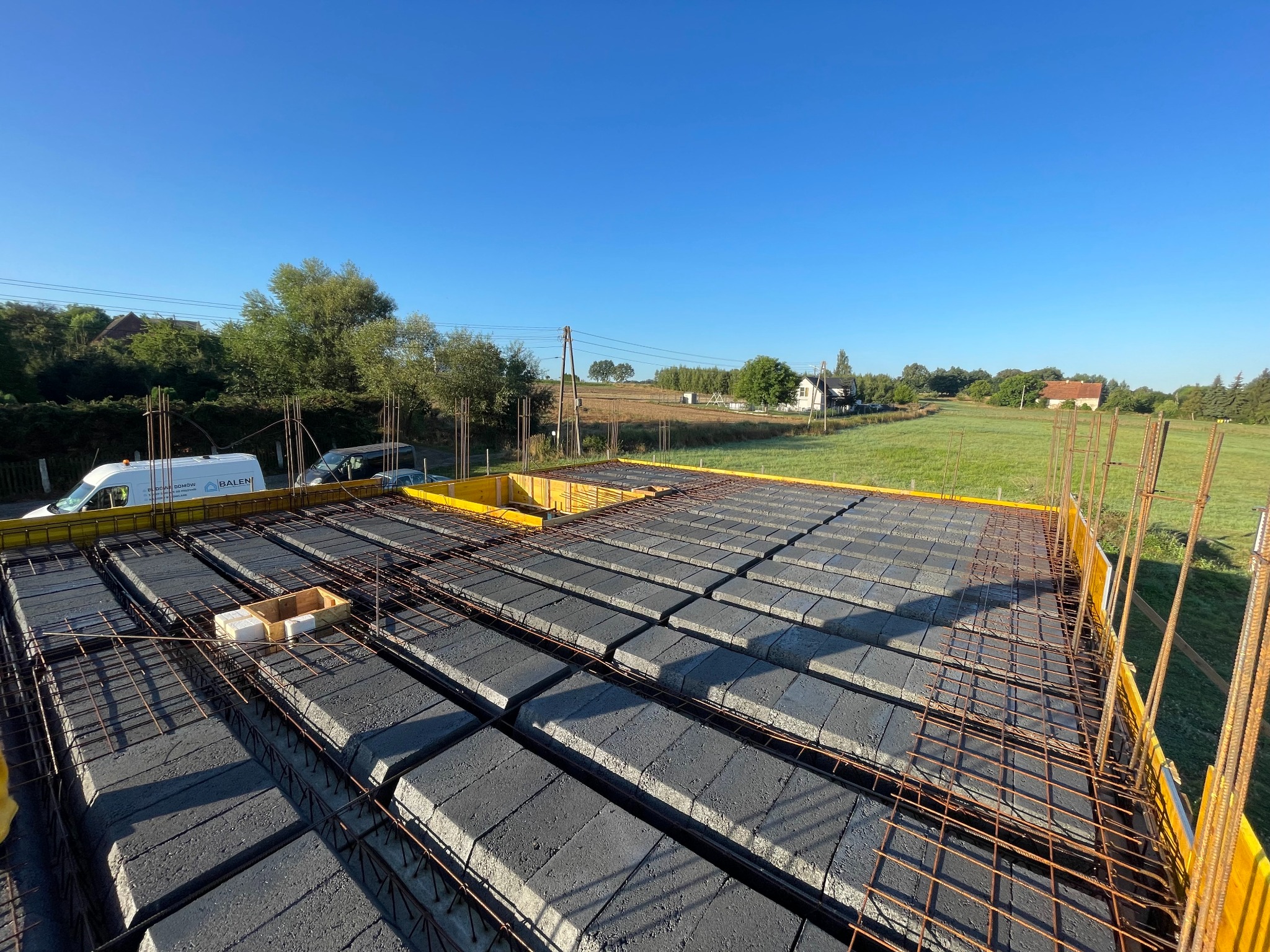 Zbrojenie stropu Teriva z szalunkiem w żółtej formie, widok z góry na tle krajobrazu z domami i błękitnym niebem. Na pierwszym planie skrzynka z elementami.