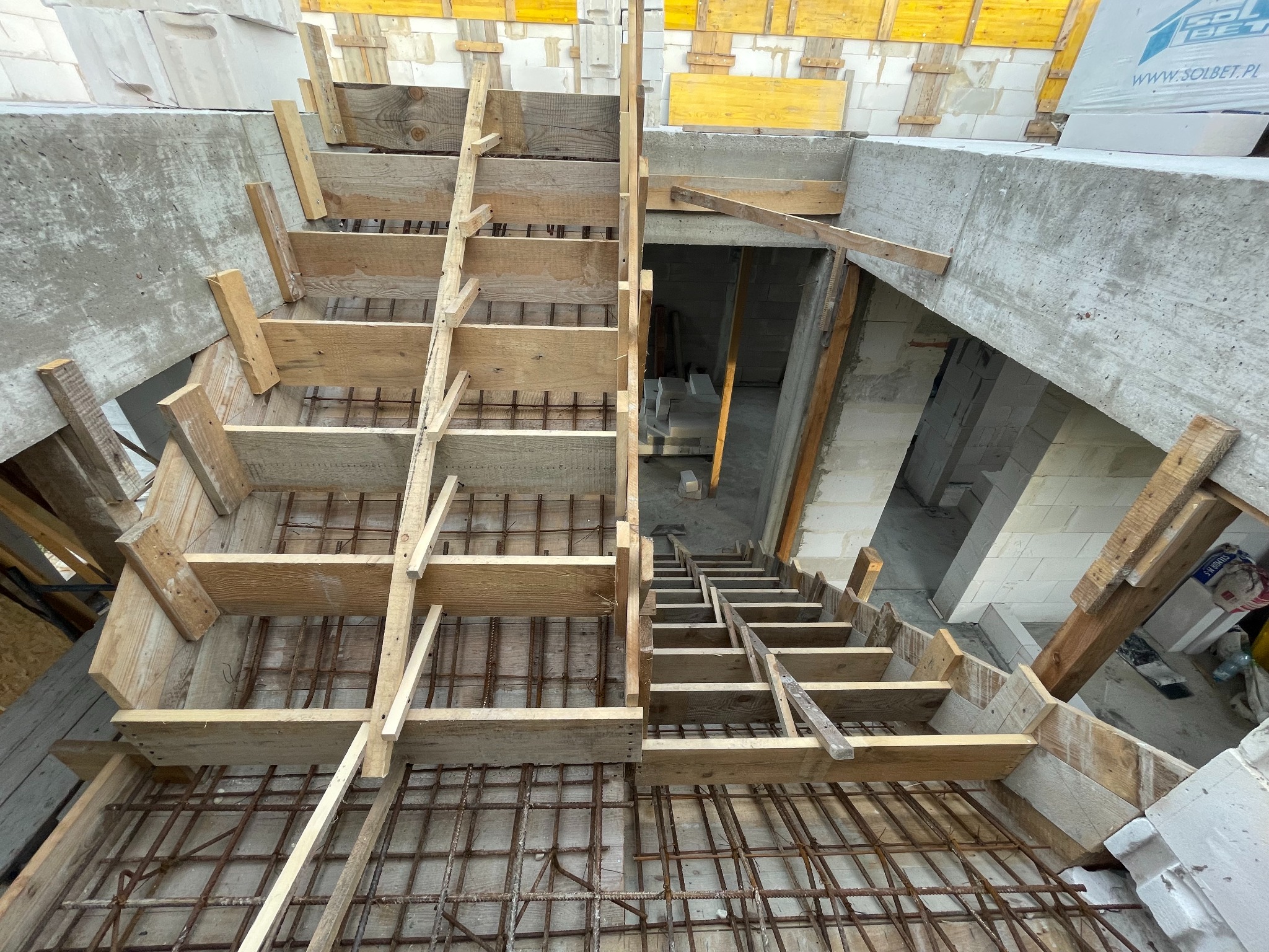 Szczegółowy widok z góry na drewniane szalunki i zbrojenie schodów betonowych w surowym stanie, z fragmentami ścian z bloczków i elementami konstrukcyjnymi w tle.