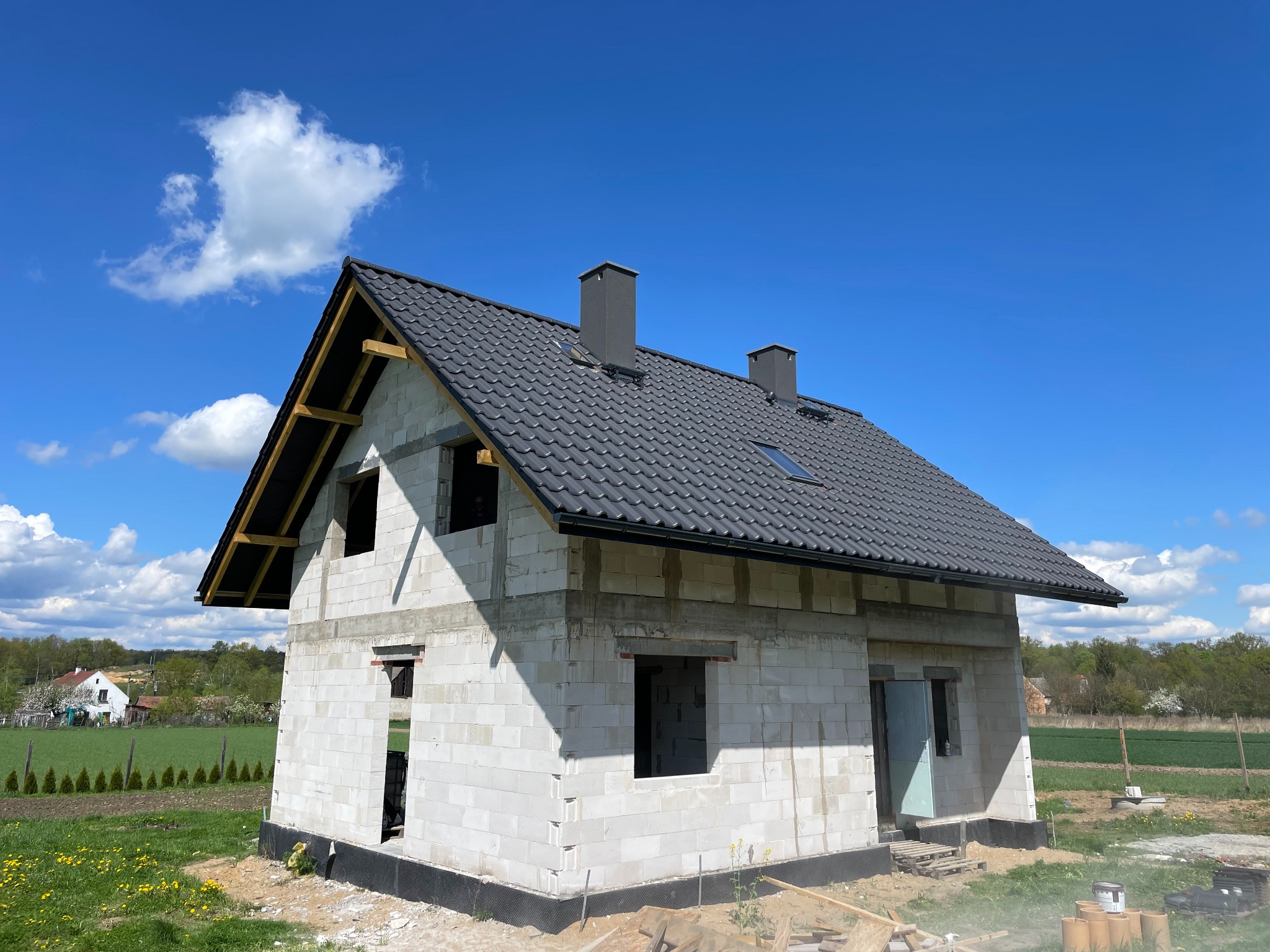 Niedokończony dom z białego betonu komórkowego z czarnym dachem, kominami i oknem dachowym. Widoczne elementy drewniane więźby dachowej. W tle błękitne niebo i zieleń.