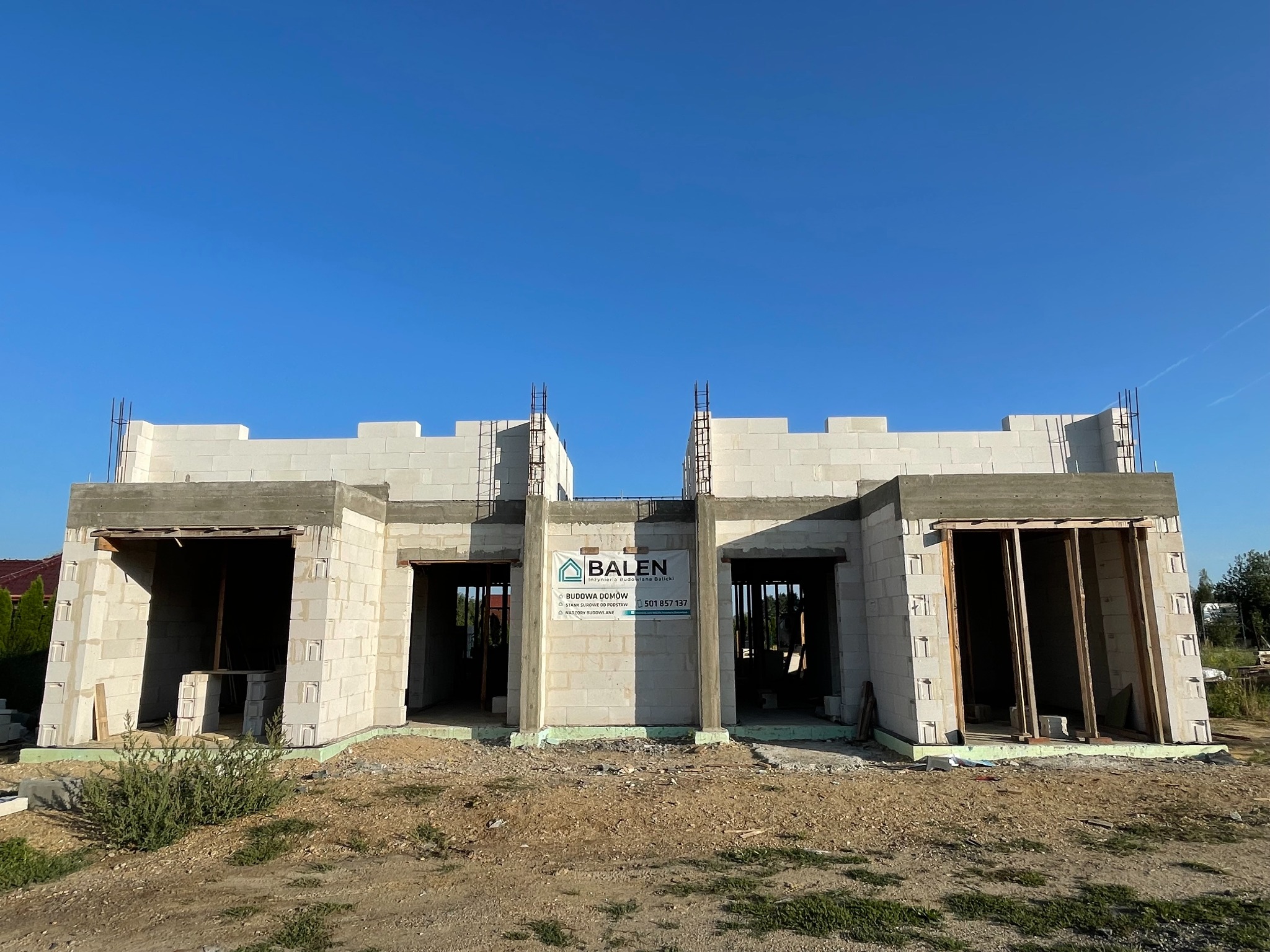 Budowa domu z białych bloczków, widoczny stan surowy otwarty, zbrojenie, logo firmy budowlanej na ścianie frontowej, błękitne niebo.