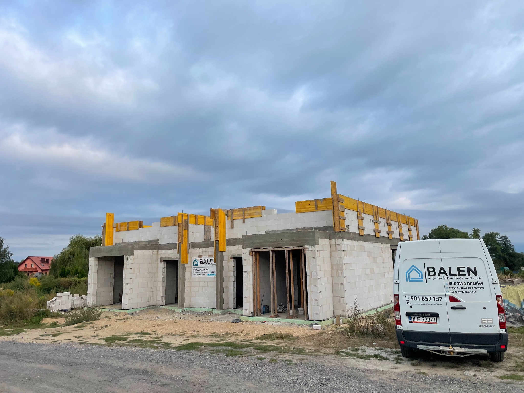 Budowa bliźniaka w stanie surowym otwartym, z widocznym szalunkiem stropu. Na pierwszym planie firmowy bus. Pochmurne niebo w tle.