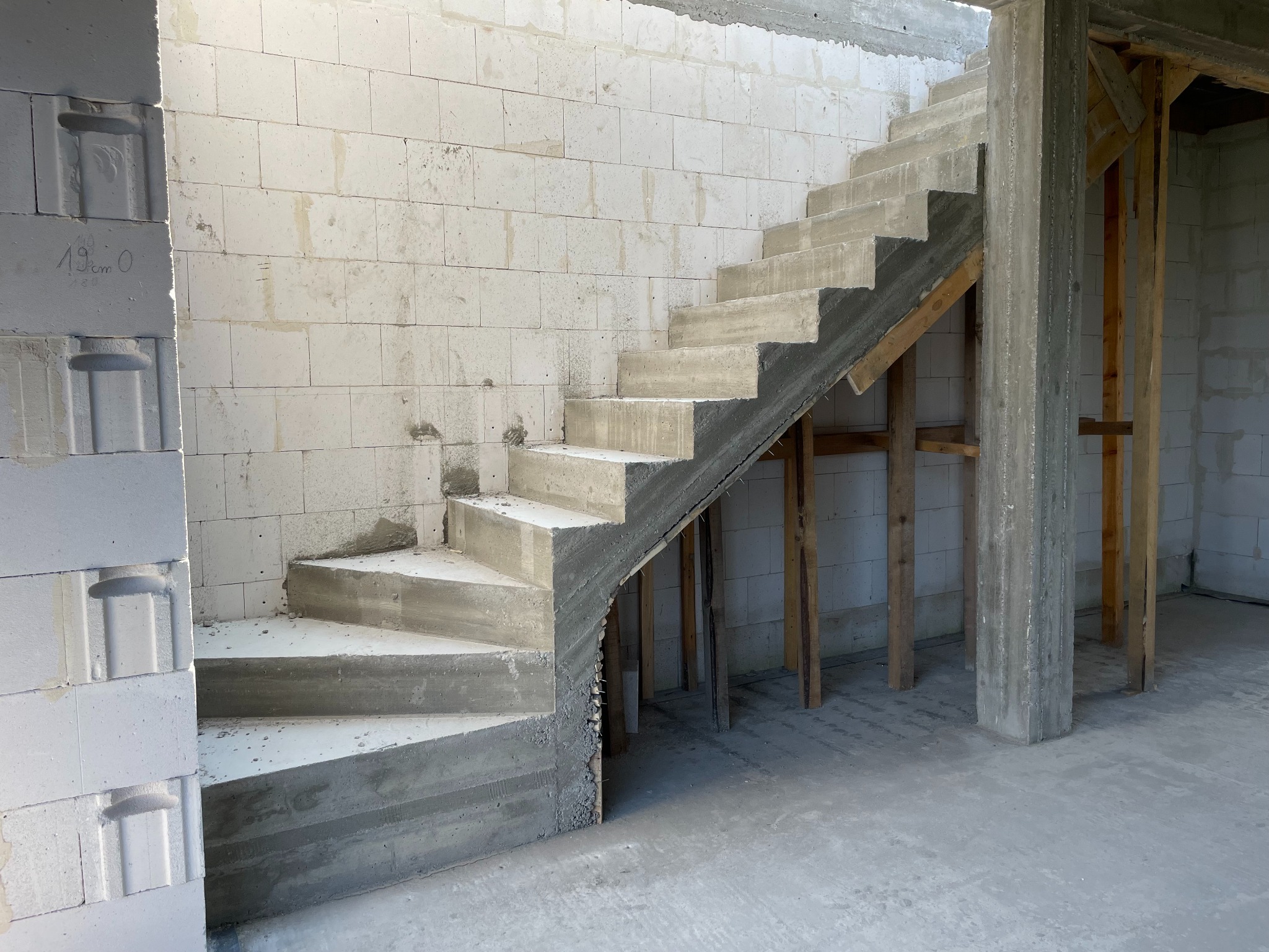 Betonowe schody w surowym stanie, wsparte na drewnianych podporach, wewnątrz budynku z białych bloczków. Widoczne niedokończone ściany i surowy beton.