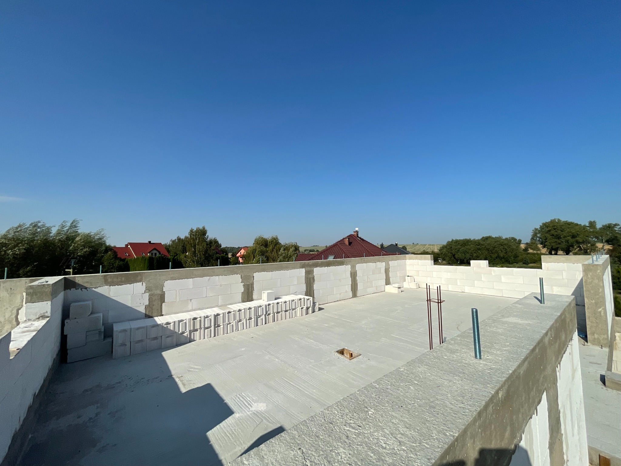 Wznoszenie ścian z betonu komórkowego na stropie budynku w trakcie budowy, widoczne zbrojenia i elementy konstrukcyjne. W tle zabudowa jednorodzinna i błękitne niebo.