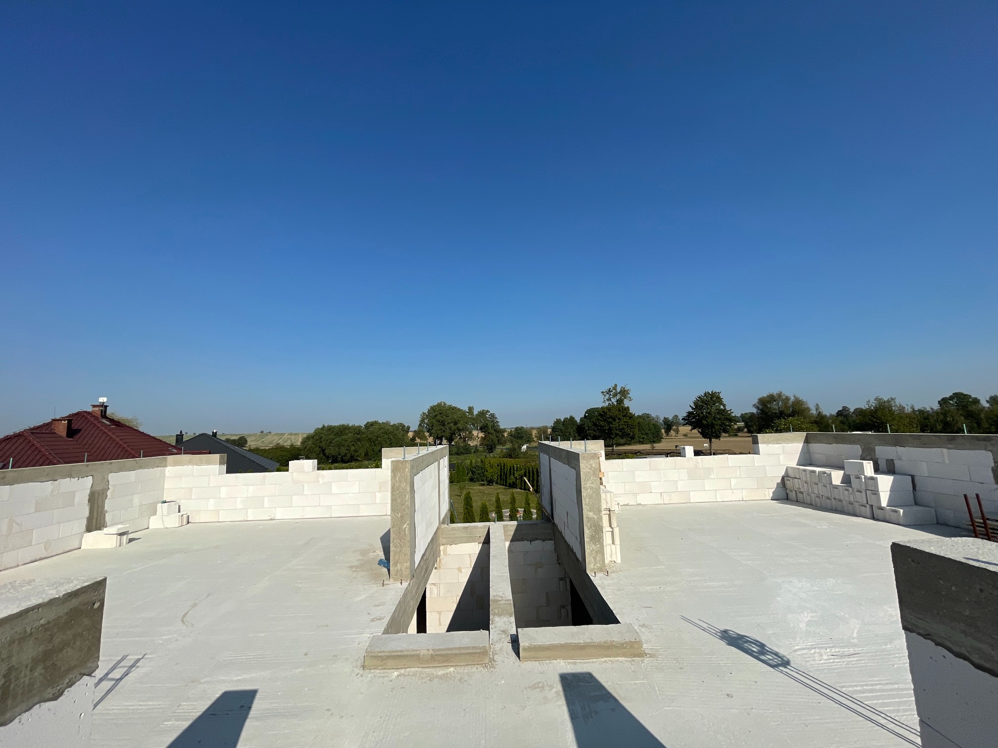 Niedokończony strop budynku z białych bloczków, z widocznymi otworami i betonowymi elementami, w tle zielony krajobraz i błękitne niebo. Budowa w toku.