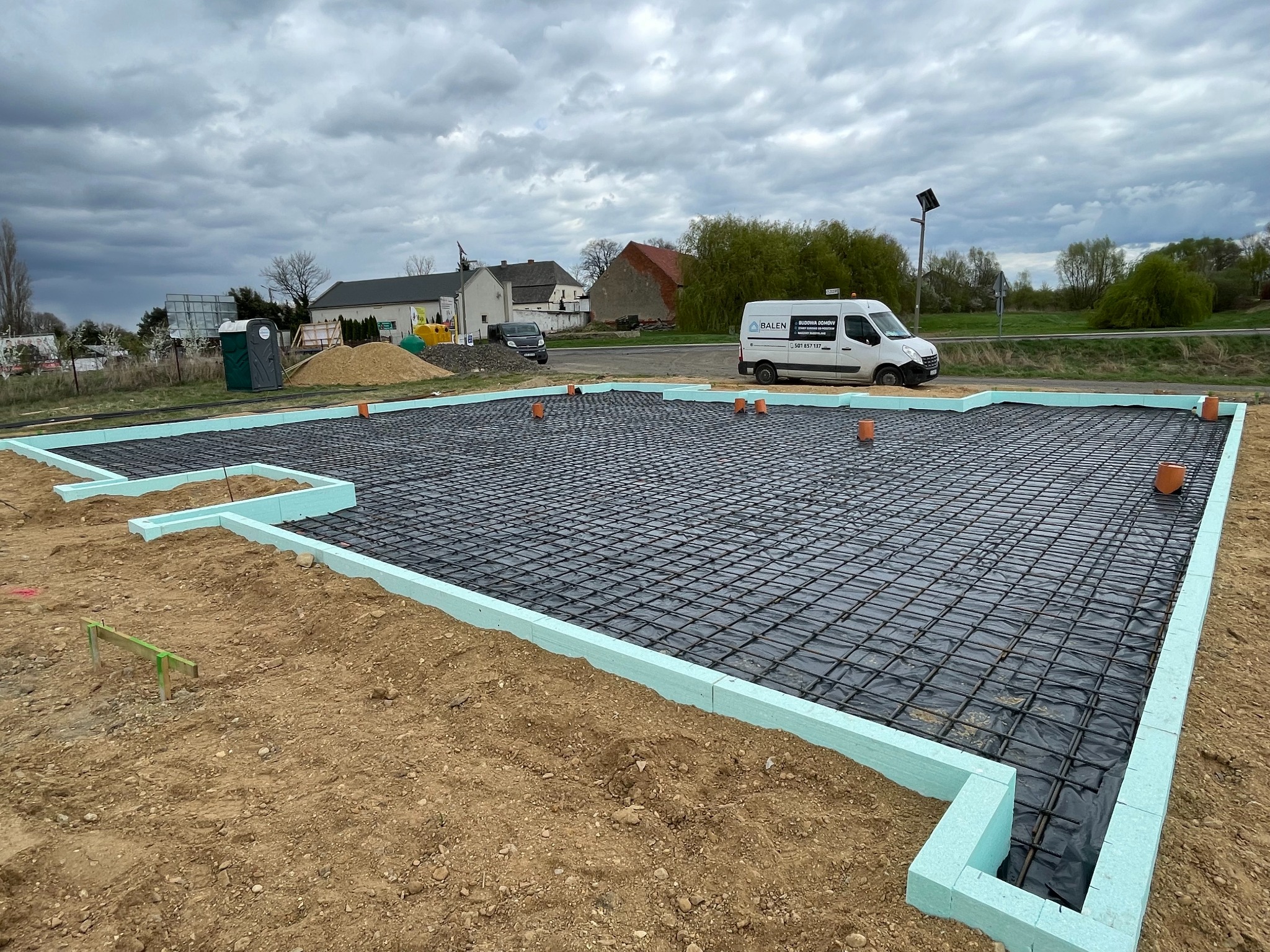 Zbrojenie fundamentów na czarnej folii, otoczone niebieskim styropianem, z widocznym samochodem firmy budowlanej i pochmurnym niebem w tle. Etap budowy domu.