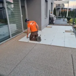 Pracownik w pomarańczowej koszulce klęczy, układając jasne płyty na zewnątrz domu, z widocznym ogrodem i częścią budynku. Płyty ułożone częściowo, widoczny etap prac.