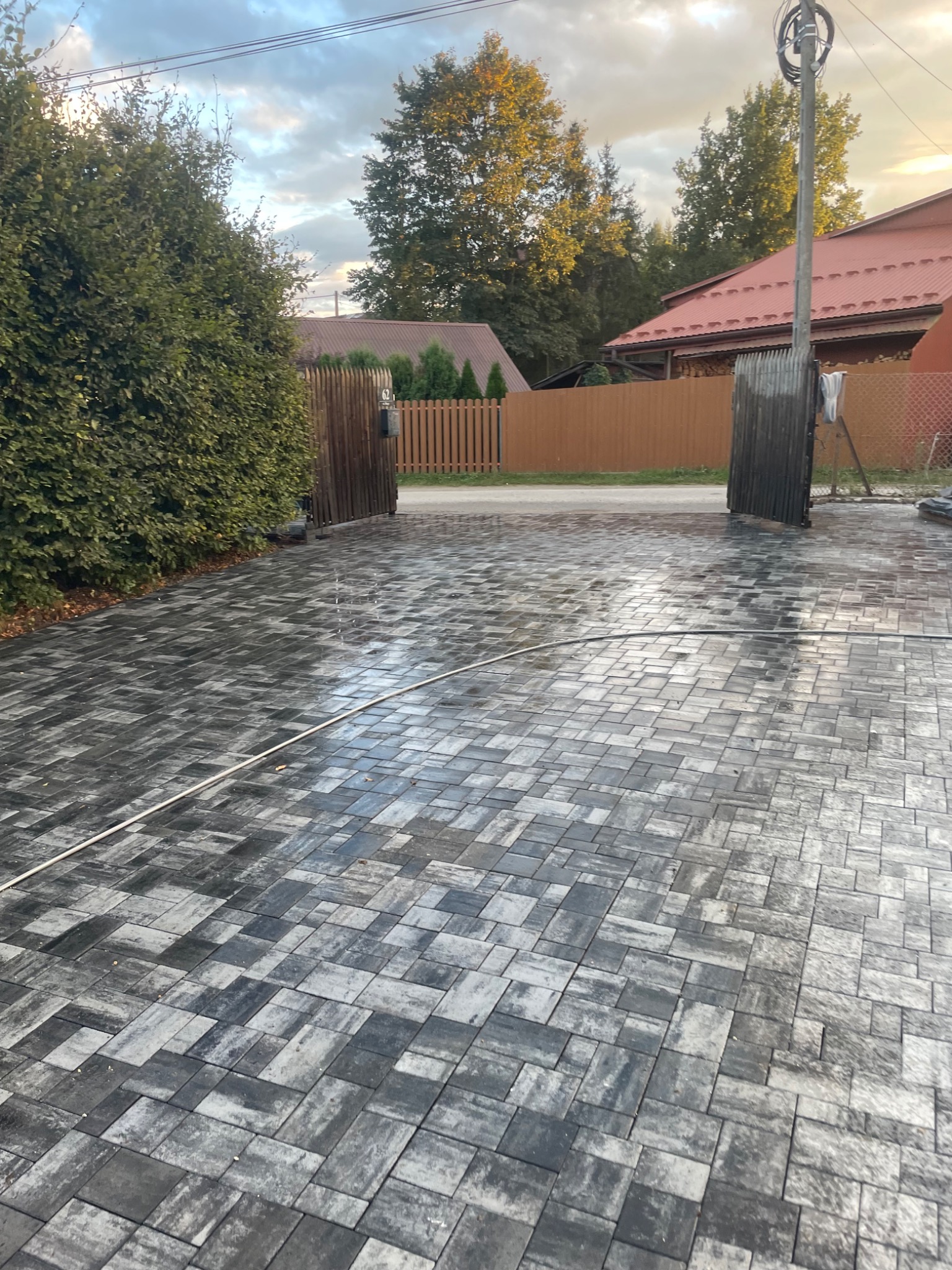 Świeżo ułożona, mokra nawierzchnia z szaro-czarnej kostki brukowej o nieregularnym wzorze, widoczne otwarte drewniane wrota w tle, zieleń i fragment domu z czerwonym dachem.