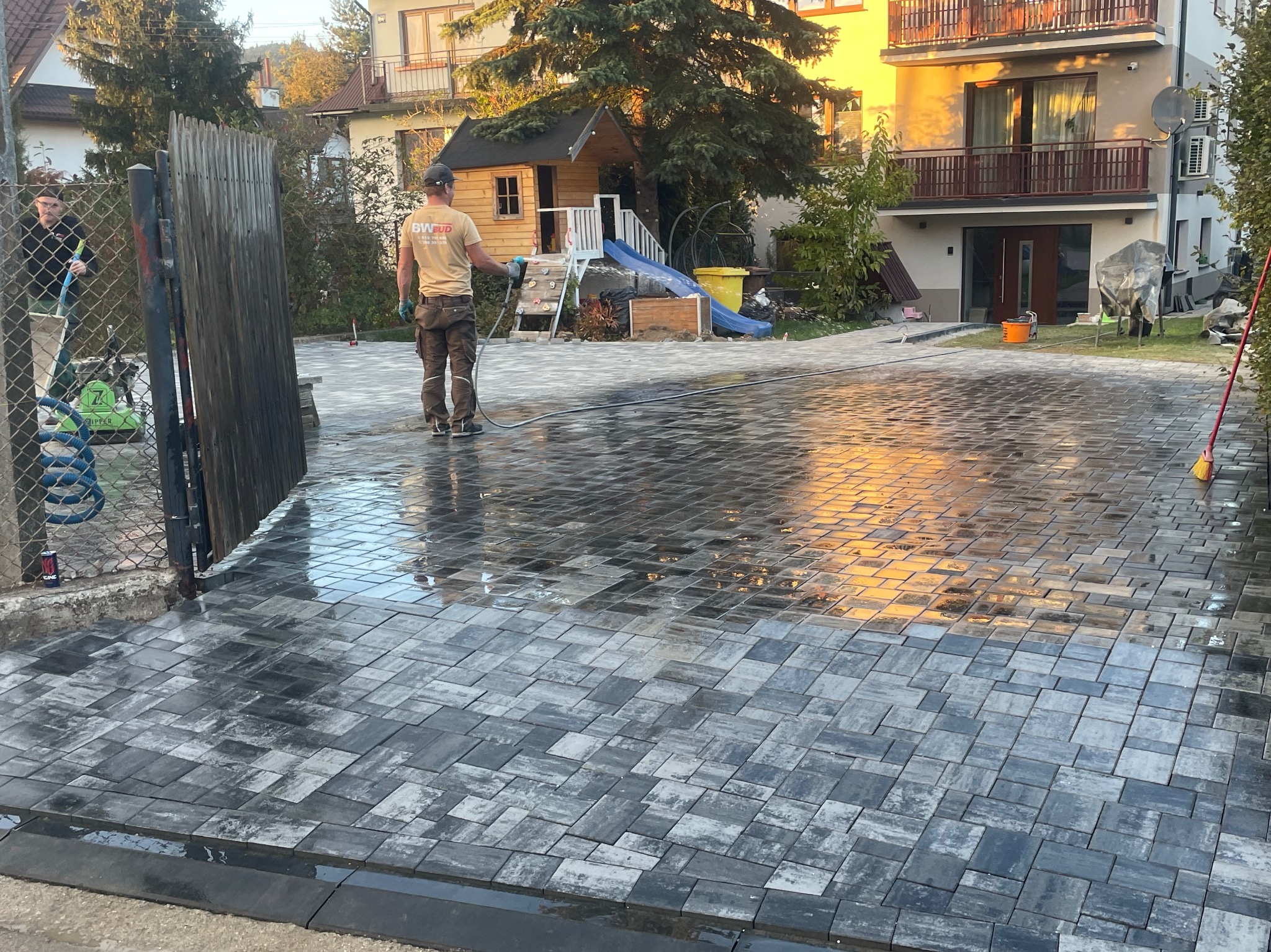 Świeżo ułożona, mokra kostka brukowa w odcieniach szarości, odbijająca promienie zachodzącego słońca, pracownik w tle spłukuje powierzchnię wodą, widoczny fragment ogrodzenia i plac zabaw dla dzieci.