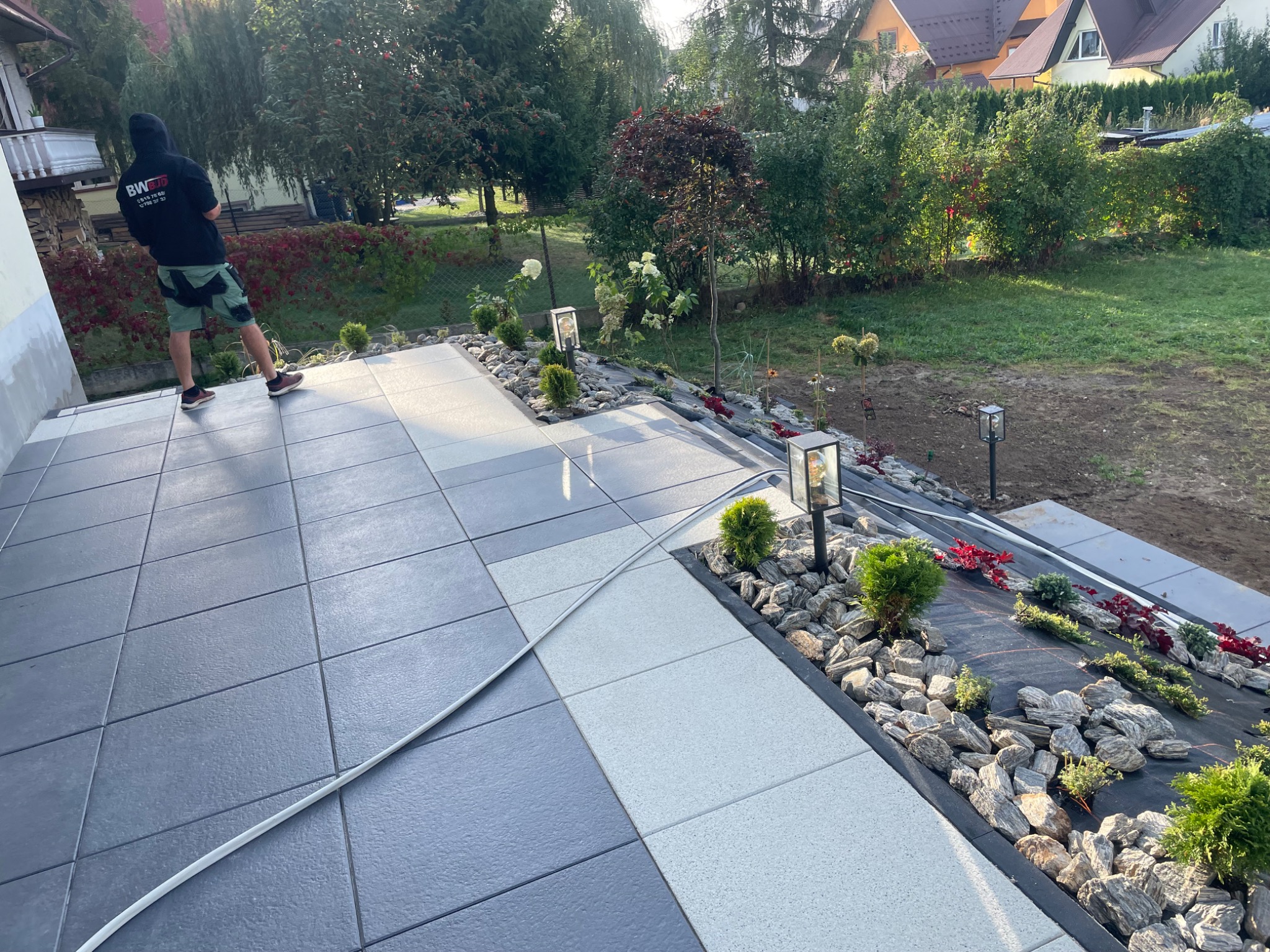 Ułożona nawierzchnia z kostki brukowej w odcieniach szarości i bieli, z ozdobnym obramowaniem z kamieni i roślin, oświetlona lampami ogrodowymi, widok z perspektywy osoby stojącej na wyższym poziomie.